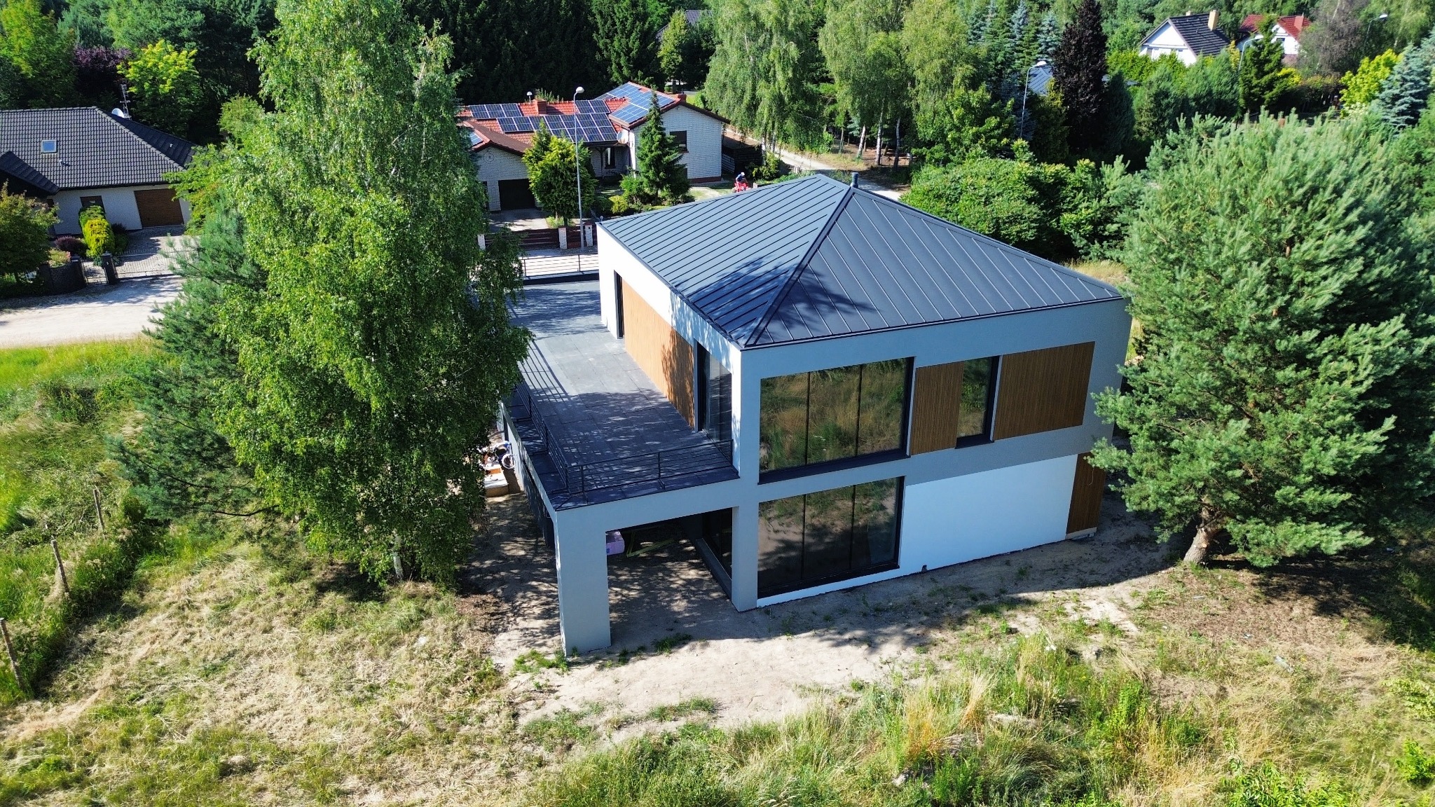 Nowoczesny, dwukondygnacyjny dom z płaskim dachem i dużymi oknami, widziany z lotu ptaka, otoczony zielenią. Minimalistyczna architektura z elementami drewna.