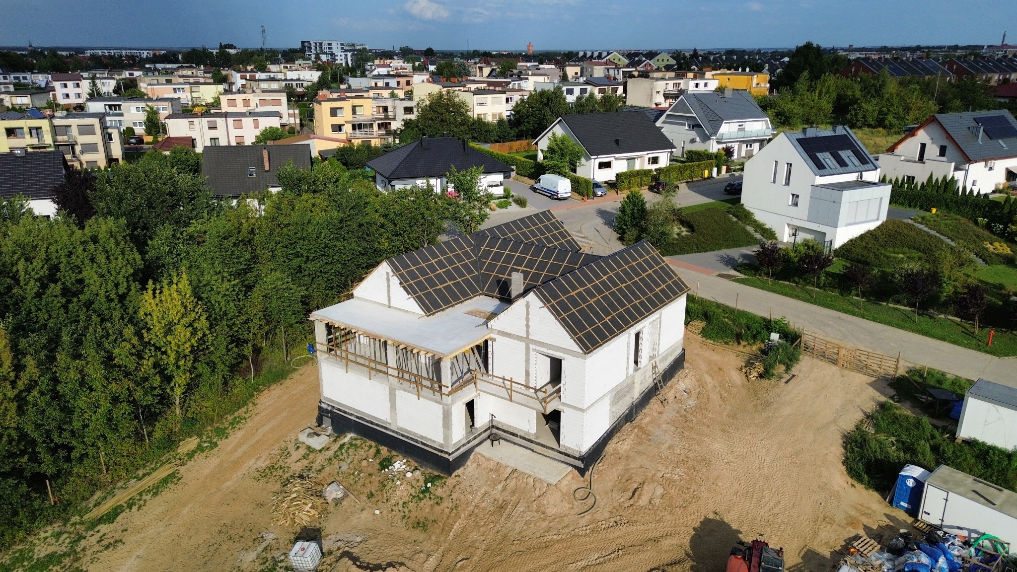 Budowa domu jednorodzinnego w trakcie realizacji. Widok z drona na budynek z białego pustaka z częściowo ułożoną konstrukcją dachu na tle zabudowy miejskiej.