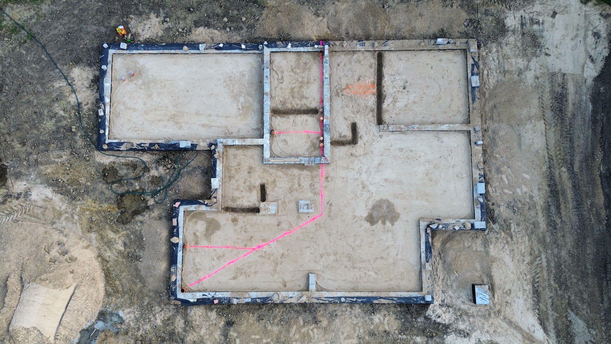 Fundamenty domu z lotu ptaka: wykopane, z izolacją, z zaznaczonymi instalacjami. Widoczne ślady opon i piasek. Ujęcie z góry, widać zarys przyszłego budynku.