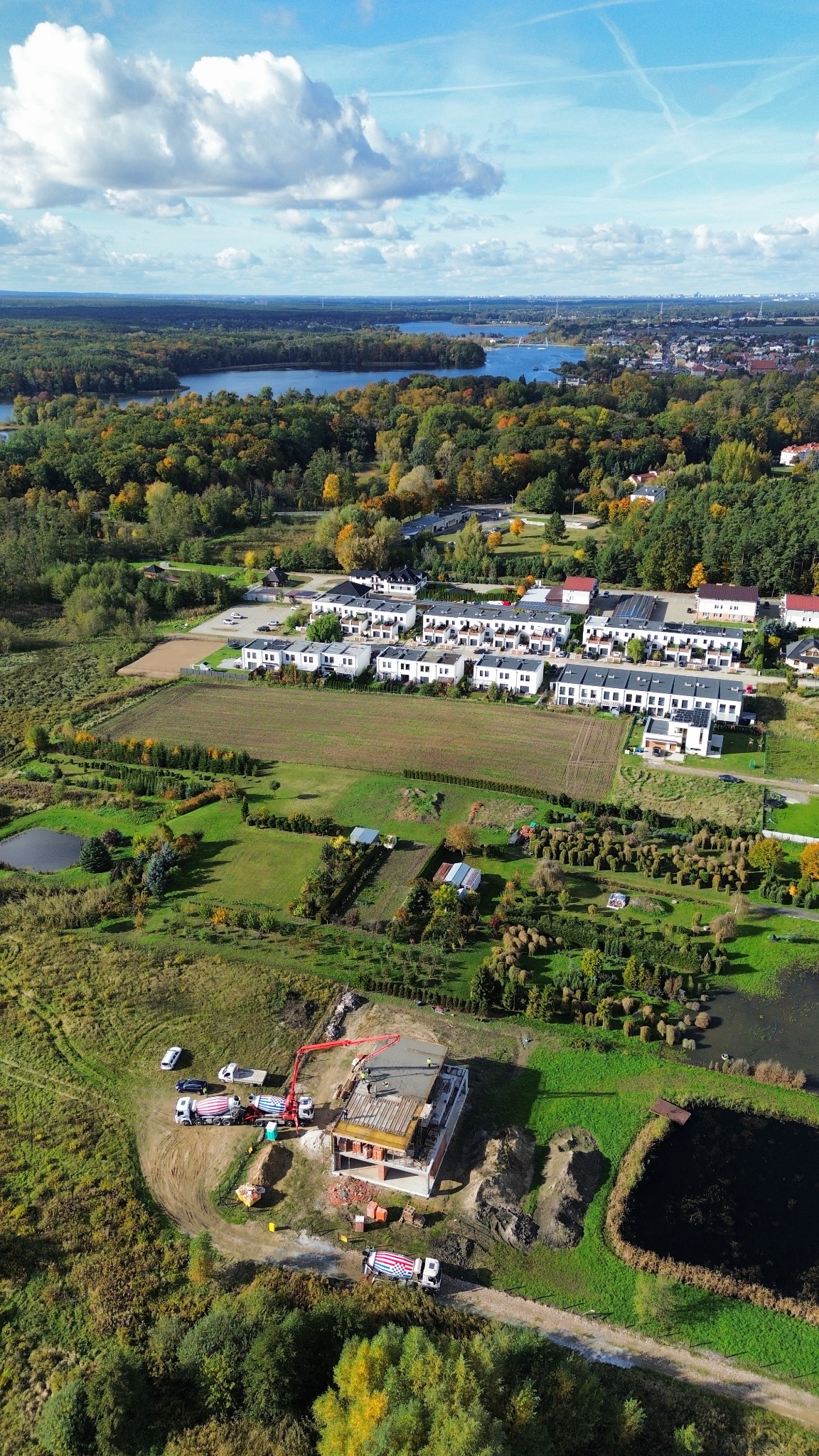 Budowa domu w otoczeniu zieleni, widok z drona. Betonowanie stropu, na budowie betoniarki i pompa do betonu. W tle zabudowa jednorodzinna i jezioro.