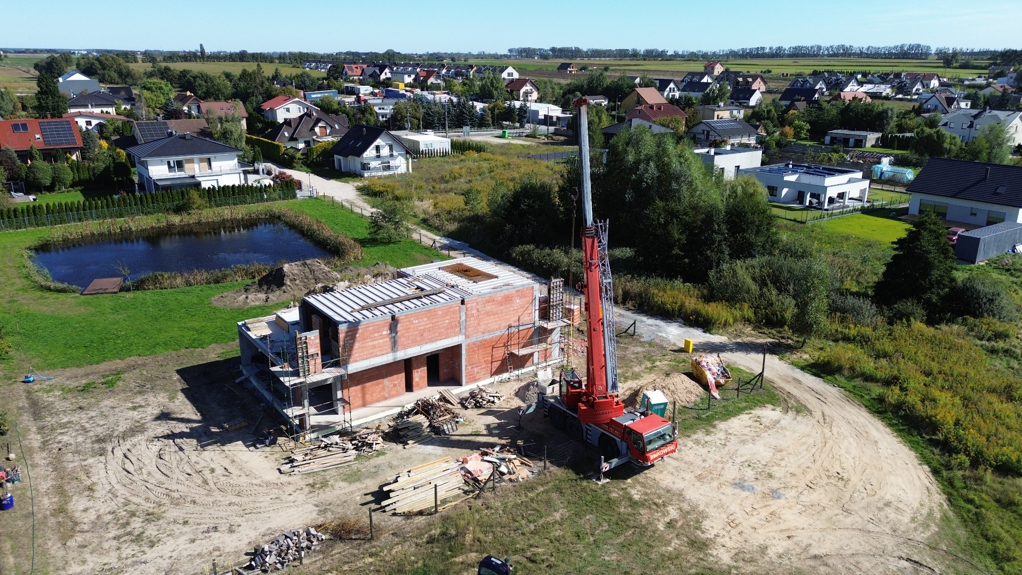 Budowa domu z czerwonej cegły, widok z góry. Na pierwszym planie dźwig, w tle zabudowa jednorodzinna i mały staw. Słoneczny dzień, niebieskie niebo.