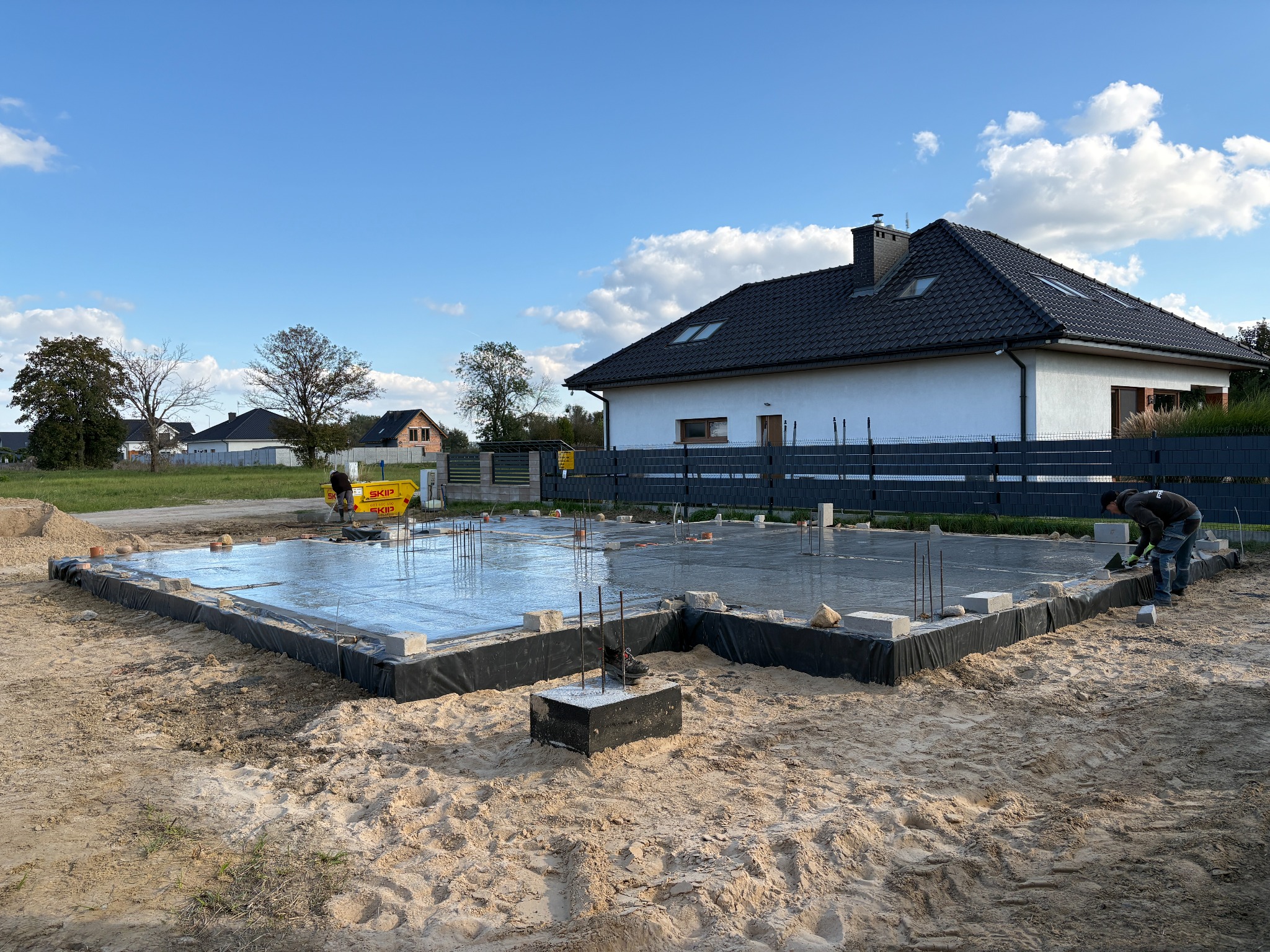 Zalane fundamenty domu jednorodzinnego z widocznym zbrojeniem i izolacją, na piaszczystym podłożu. W tle dom w trakcie budowy i błękitne niebo.