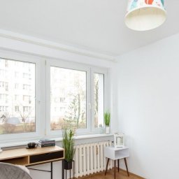 Martin Krawiec - Jasne wnętrze pokoju z biurkiem, krzesłem i oknem, ściany pomalowane na biało, widoczny grzejnik i dekoracje.