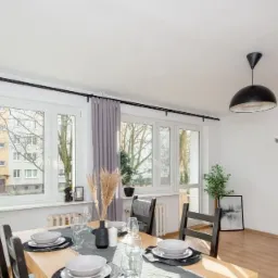 Jasna jadalnia z oknem na blok, drewniany stół nakryty dla kilku osób, czarne krzesła i lampa, szare zasłony i dekoracje roślinne