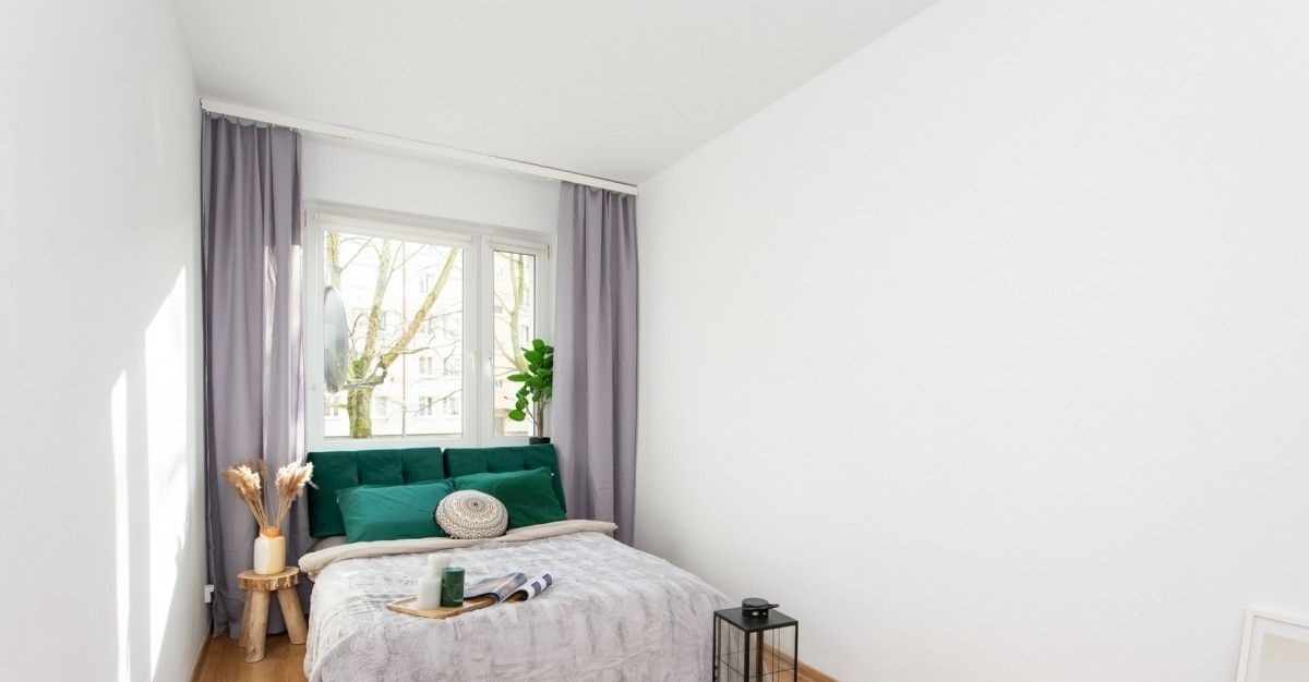 Jasne wnętrze sypialni po odświeżeniu z łóżkiem z zielonym zagłówkiem, szarymi zasłonami i drewnianym stolikiem nocnym z dekoracjami.