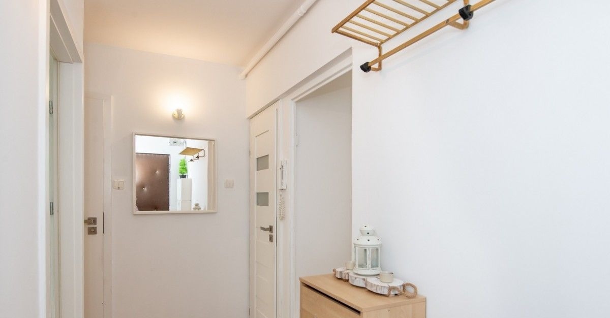 Jasne wnętrze przedpokoju z białymi ścianami, drewnianą komodą z dekoracyjną latarenką i lustrem odbijającym fragment innego pomieszczenia.