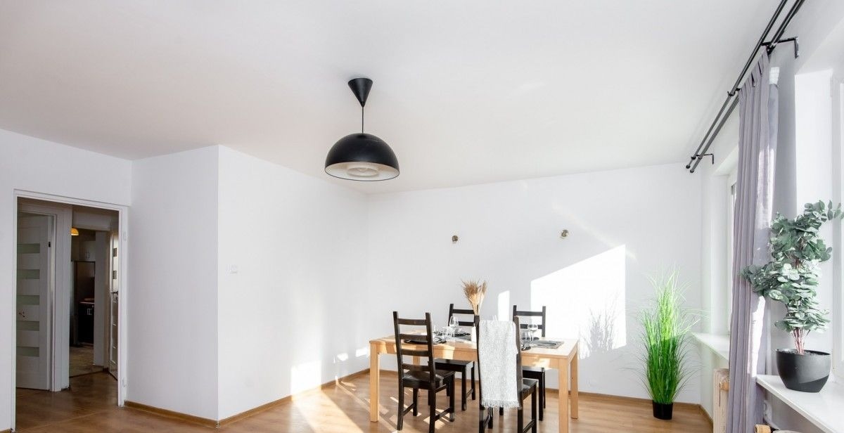 Jasne wnętrze mieszkania z drewnianą podłogą, białymi ścianami i sufitem, oświetlone czarną lampą wiszącą nad drewnianym stołem z czarnymi krzesłami, dekorowane roślinami w doniczkach i zbożem...