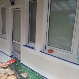 ROM Sławomir - Biała elewacja budynku po malowaniu, okna zabezpieczone folią i taśmą malarską. Widoczne detale: lampa zewnętrzna, taśma malarska, folia ochronna na podłożu.