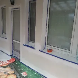 Biała elewacja budynku po malowaniu, okna zabezpieczone folią i taśmą malarską. Widoczne detale: lampa zewnętrzna, taśma malarska, folia ochronna na podłożu.