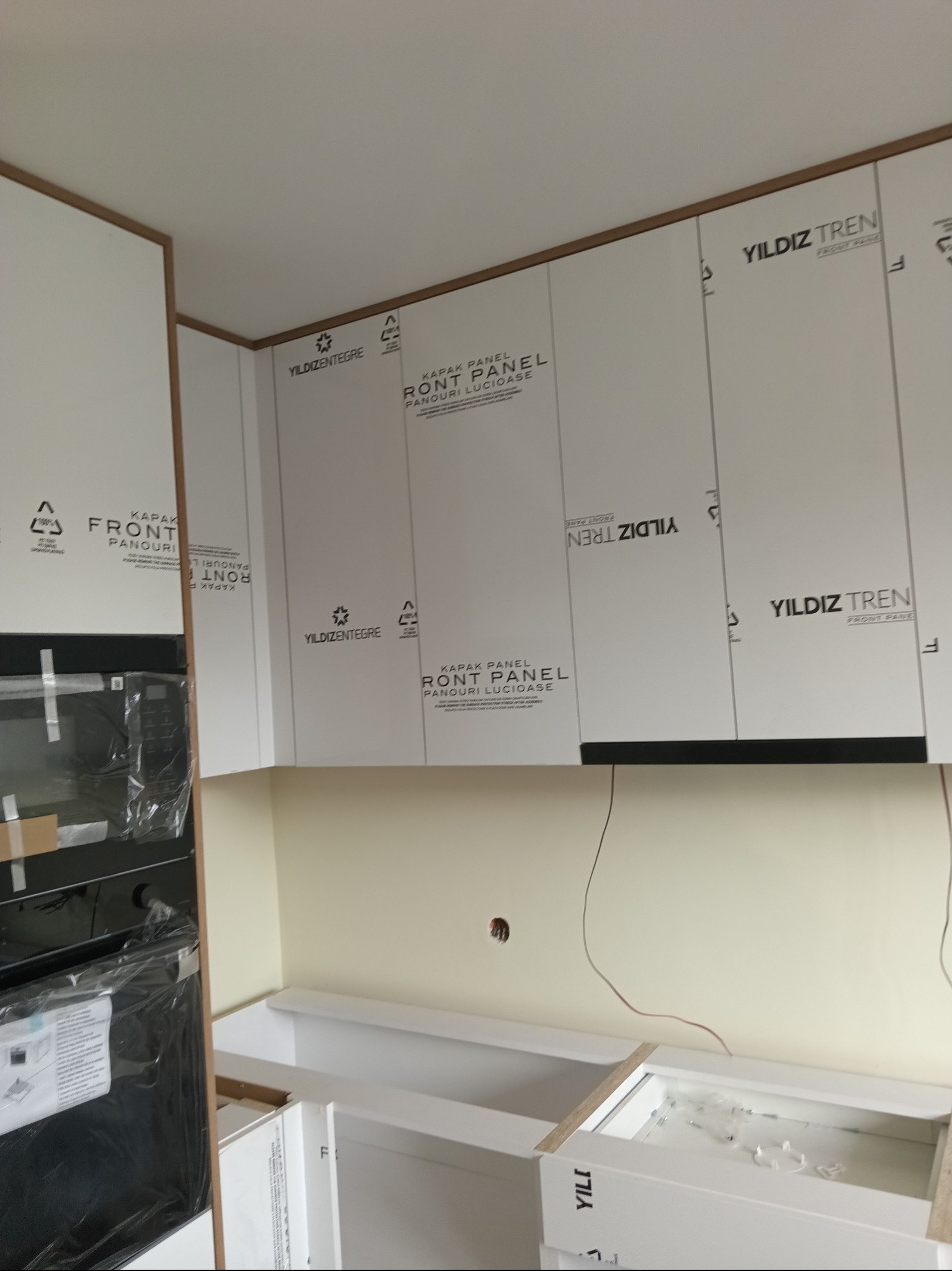 Montaż białych szafek kuchennych z widocznymi etykietami producenta na frontach. Widoczny fragment piekarnika w folii ochronnej i niedokończone dolne szafki.