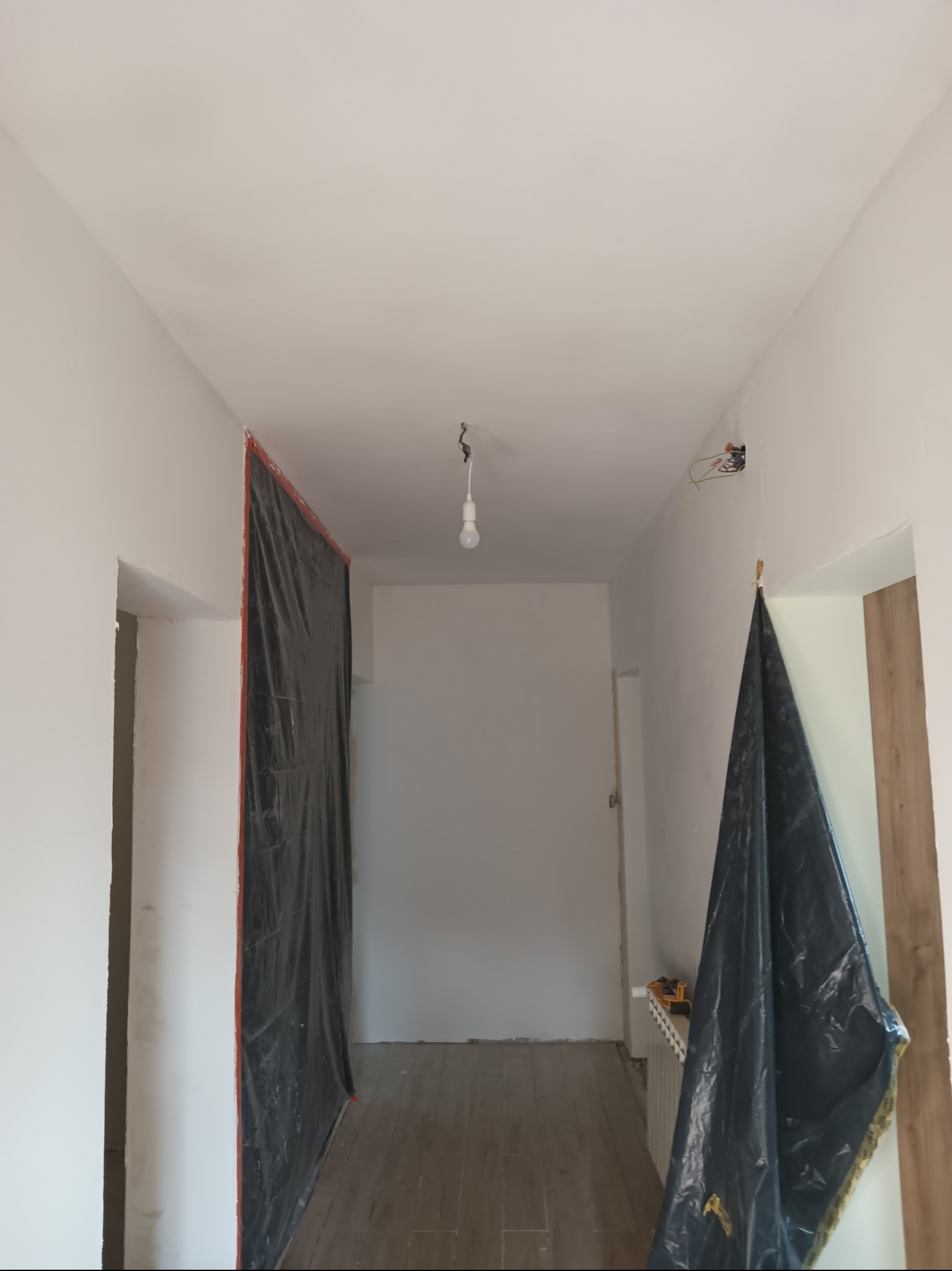Korytarz w trakcie malowania: ściany przygotowane, zabezpieczone folią malarską, widoczne narzędzia i niedokończone gniazdko elektryczne.