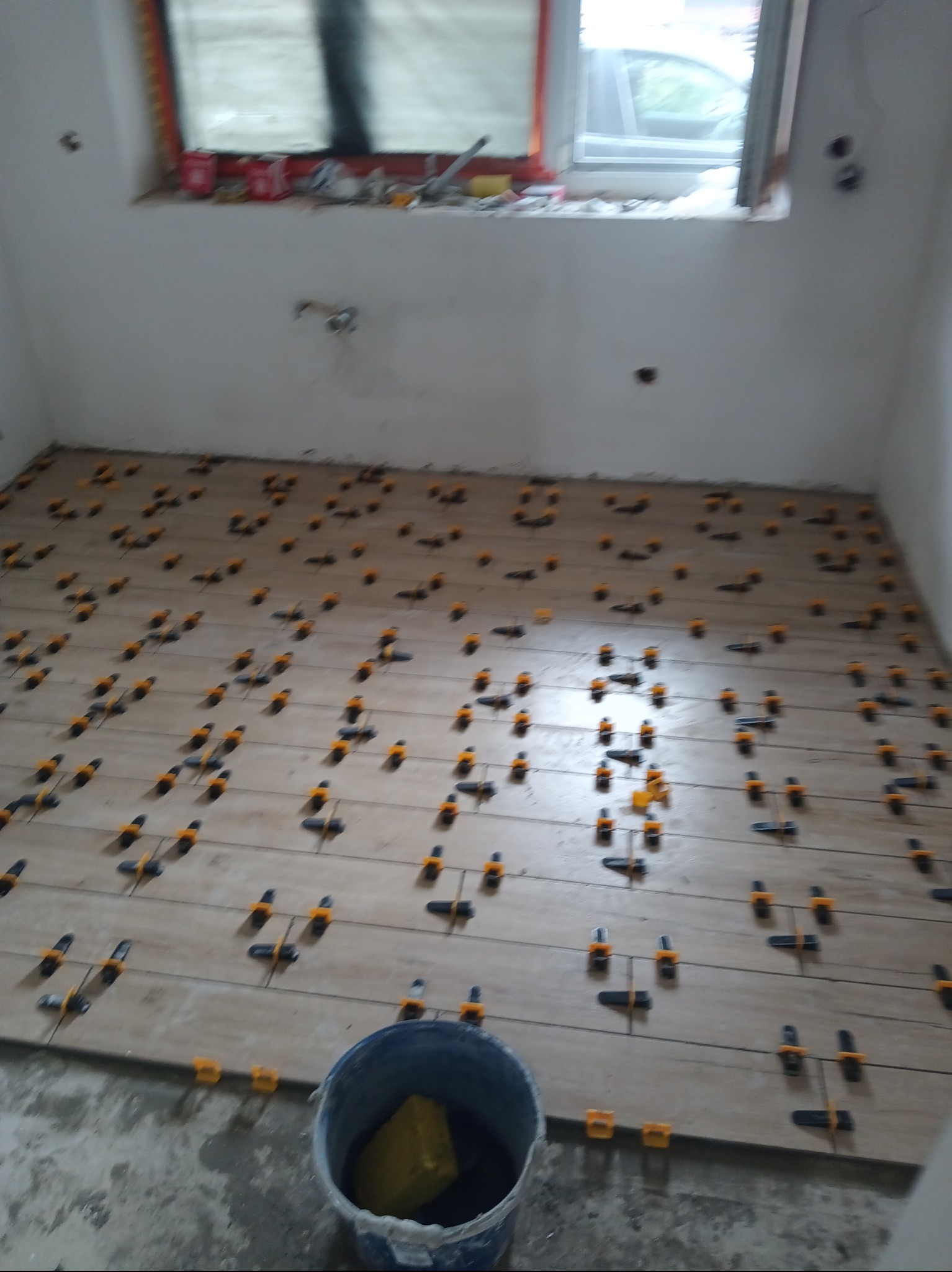 Układanie płytek podłogowych w jasnym odcieniu, z użyciem żółto-czarnych klipsów poziomujących. W tle okno z widocznymi narzędziami i pusty kubeł na pierwszym planie.