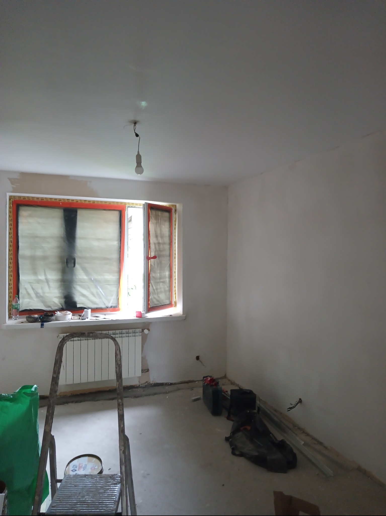 Pokój w trakcie remontu: zabezpieczone okno folią, ściany po tynkowaniu, widoczna drabina, narzędzia i materiały malarskie. Przygotowanie do malowania ścian.