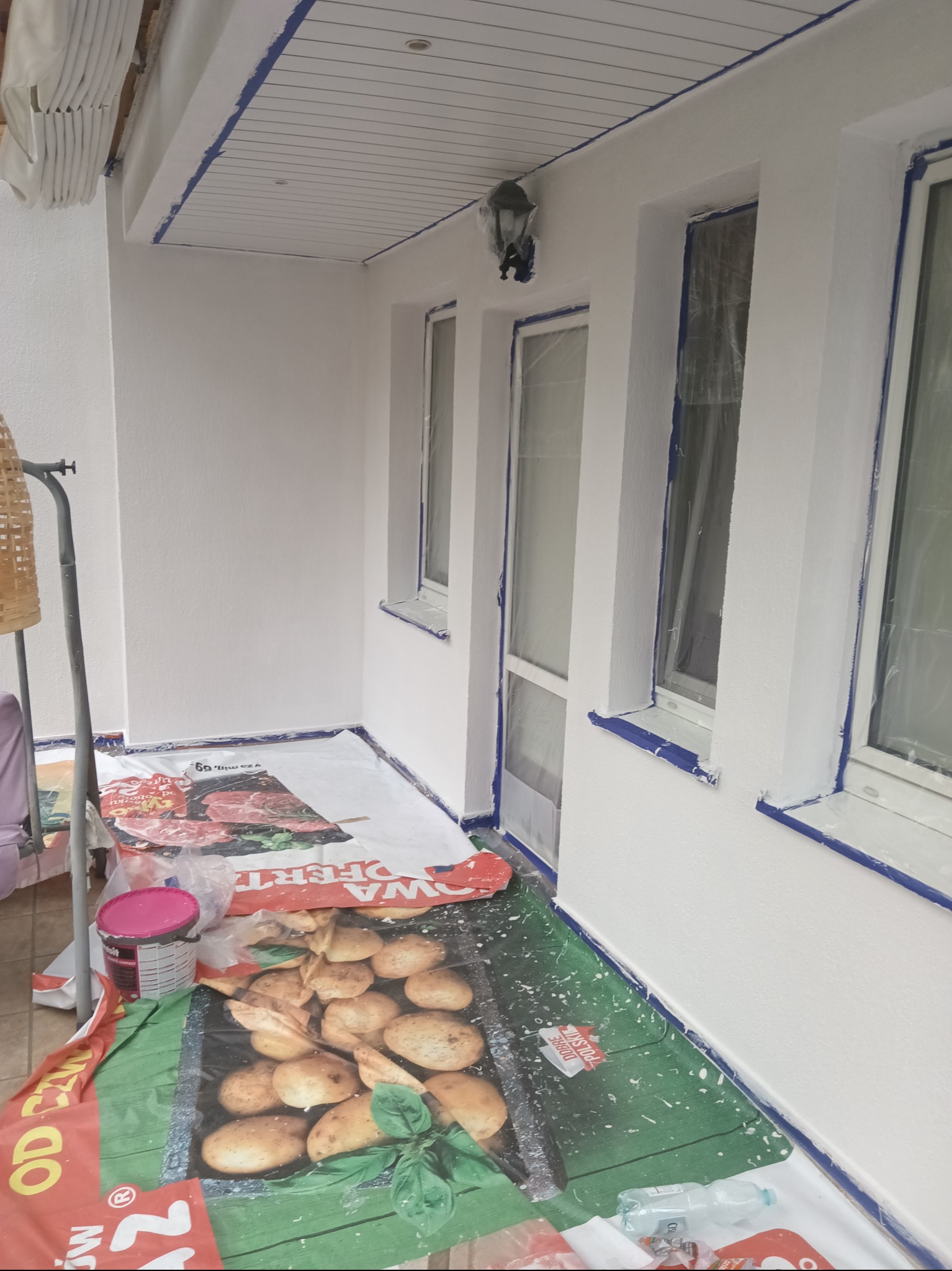 Białe ściany po malowaniu na balkonie, zabezpieczone okna folią, podłoga przykryta folią malarską. Widoczna puszka z farbą i lampa na ścianie.