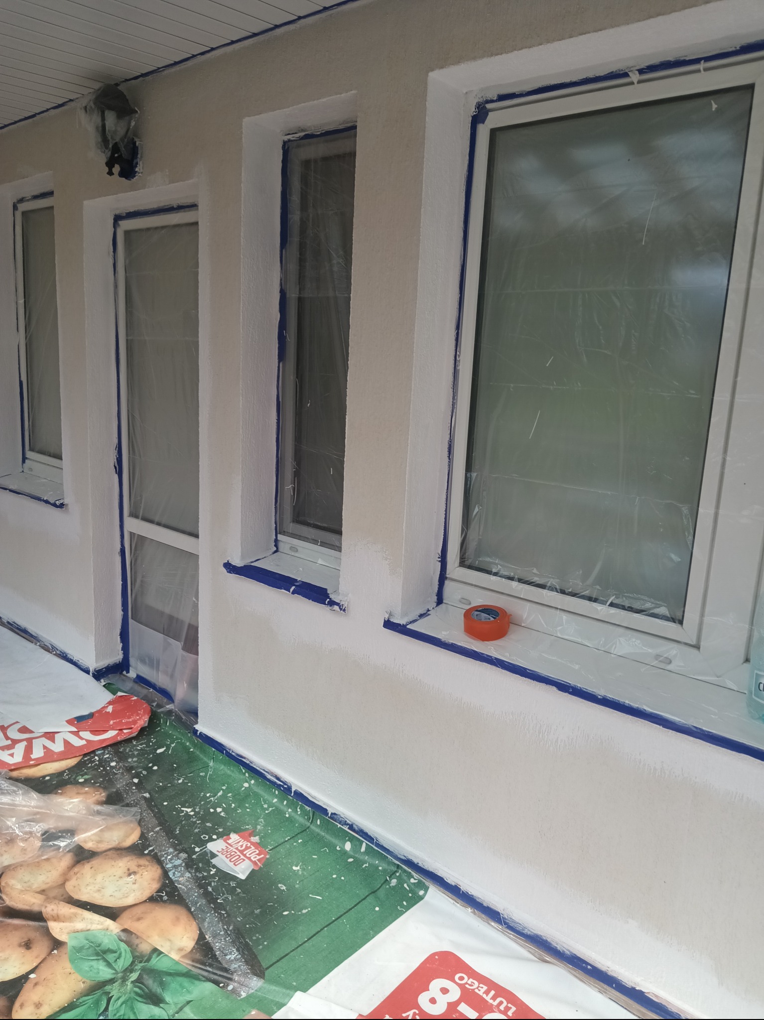 Biała elewacja budynku po malowaniu, okna zabezpieczone folią i taśmą malarską. Widoczne detale: lampa zewnętrzna, taśma malarska, folia ochronna na podłożu.