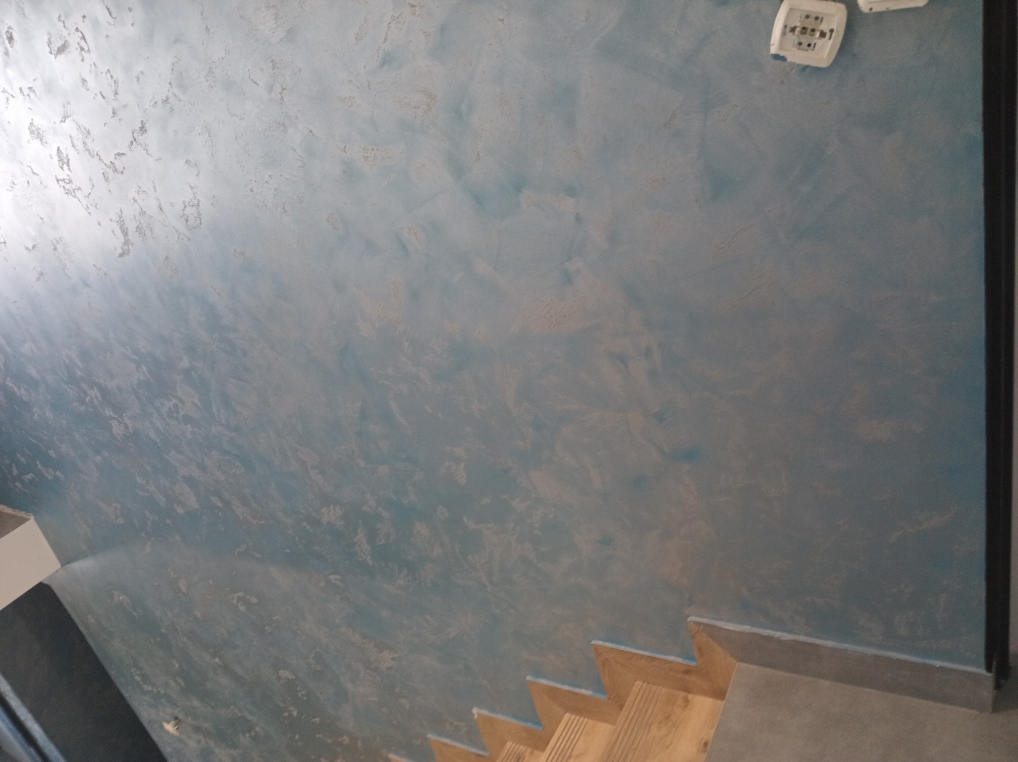 Błękitna ściana z efektem dekoracyjnym, widoczne schody drewniane i białe gniazdka elektryczne. Nietypowy kąt ujęcia podkreśla fakturę ściany.