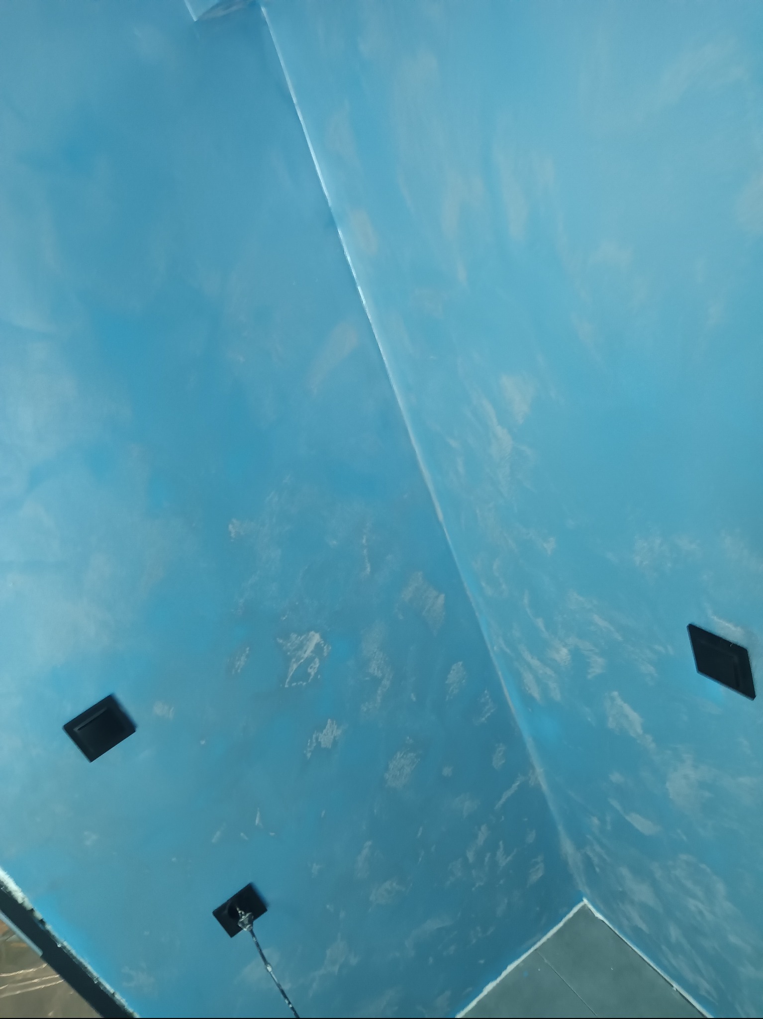 Błękitne ściany z efektem chmur w Radomiu, wykończone czarnymi oprawami oświetleniowymi. Dekoracyjne malowanie wnętrz, widok z dołu, podkreślający unikalną fakturę.