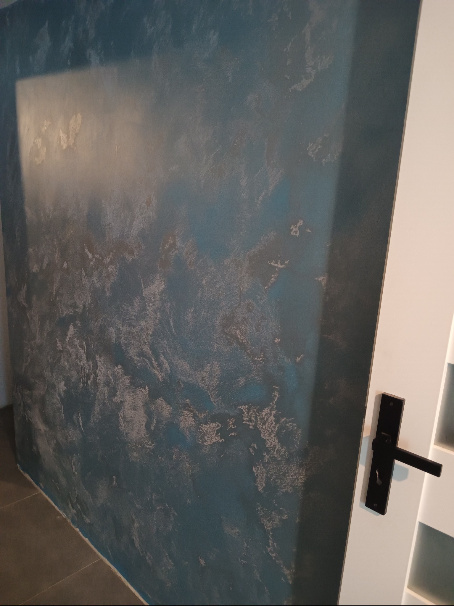 Dekoracyjna ściana w odcieniach błękitu i szarości z efektem chmur, widoczna obok białych drzwi z czarną klamką. Unikalna faktura i wzór na ścianie.