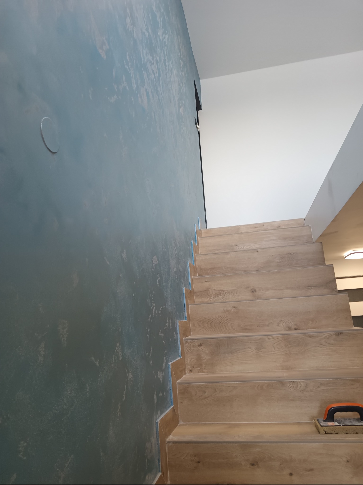 Stylowe schody wyłożone panelami imitującymi drewno, z widoczną ścianą o dekoracyjnym, ciemnym wykończeniu. Na schodach leży gąbka do czyszczenia.