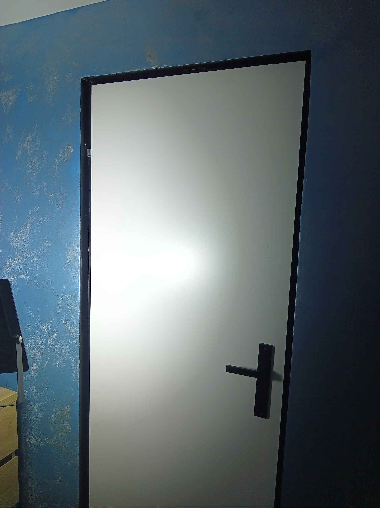 Białe drzwi z czarną ramą i klamką na tle ściany pomalowanej na niebiesko z efektem dekoracyjnym. Widoczny fragment mebla.