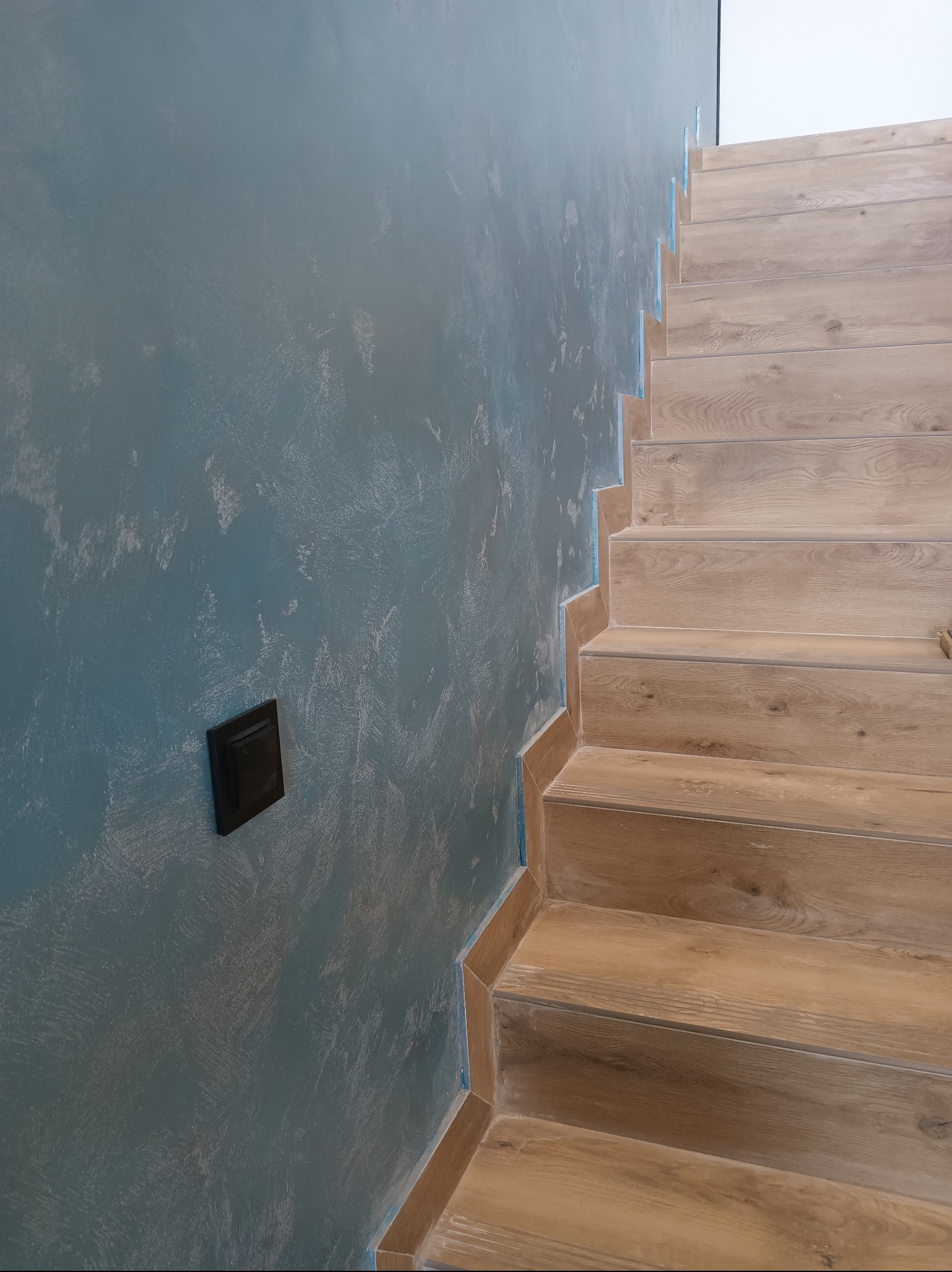 Ściana z dekoracyjnym, niebieskim tynkiem strukturalnym i drewnopodobne schody z wykończonymi krawędziami. Widoczny czarny włącznik światła na ścianie.