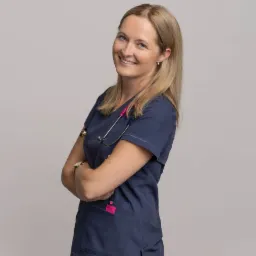 Uśmiechnięta blondynka w granatowym uniformie medycznym ze stetoskopem z różowymi elementami, pozująca na neutralnym tle.