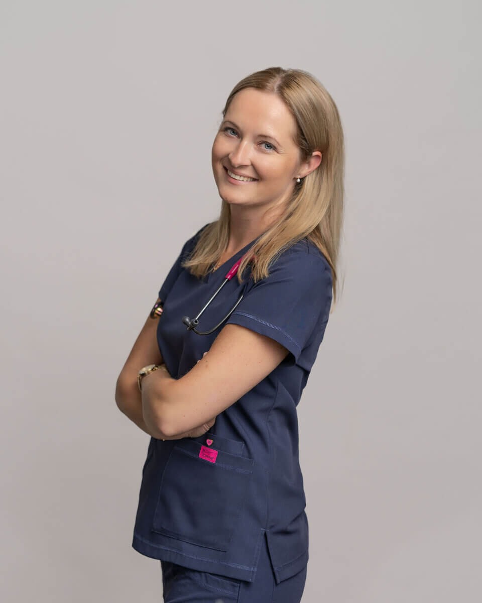 Uśmiechnięta blondynka w granatowym uniformie medycznym ze stetoskopem z różowymi elementami, pozująca na neutralnym tle.