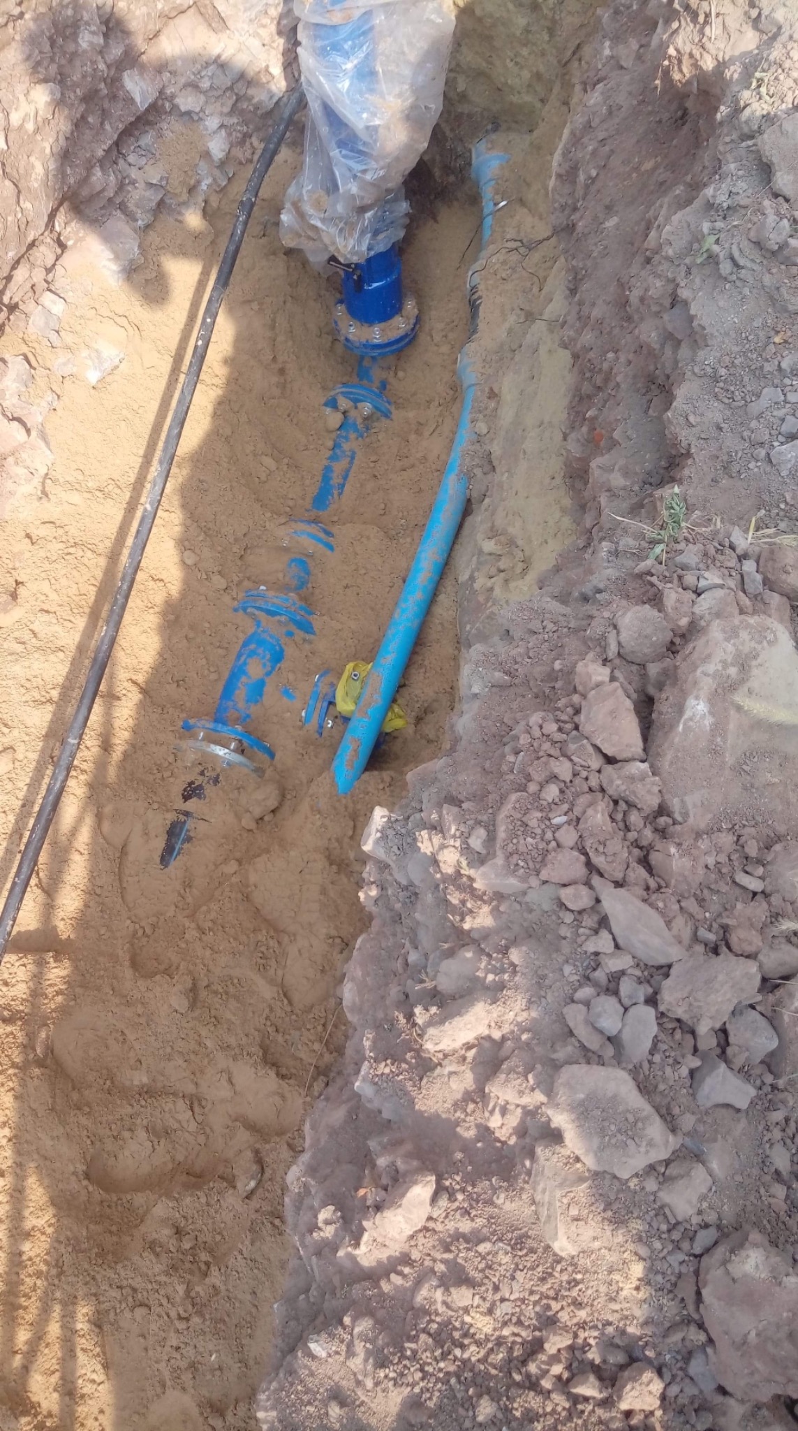 Wykop z widocznymi niebieskimi rurami wodociągowymi z zaworami, złączkami kołnierzowymi i elementami zabezpieczonymi folią, ułożonymi w piaszczystym gruncie.