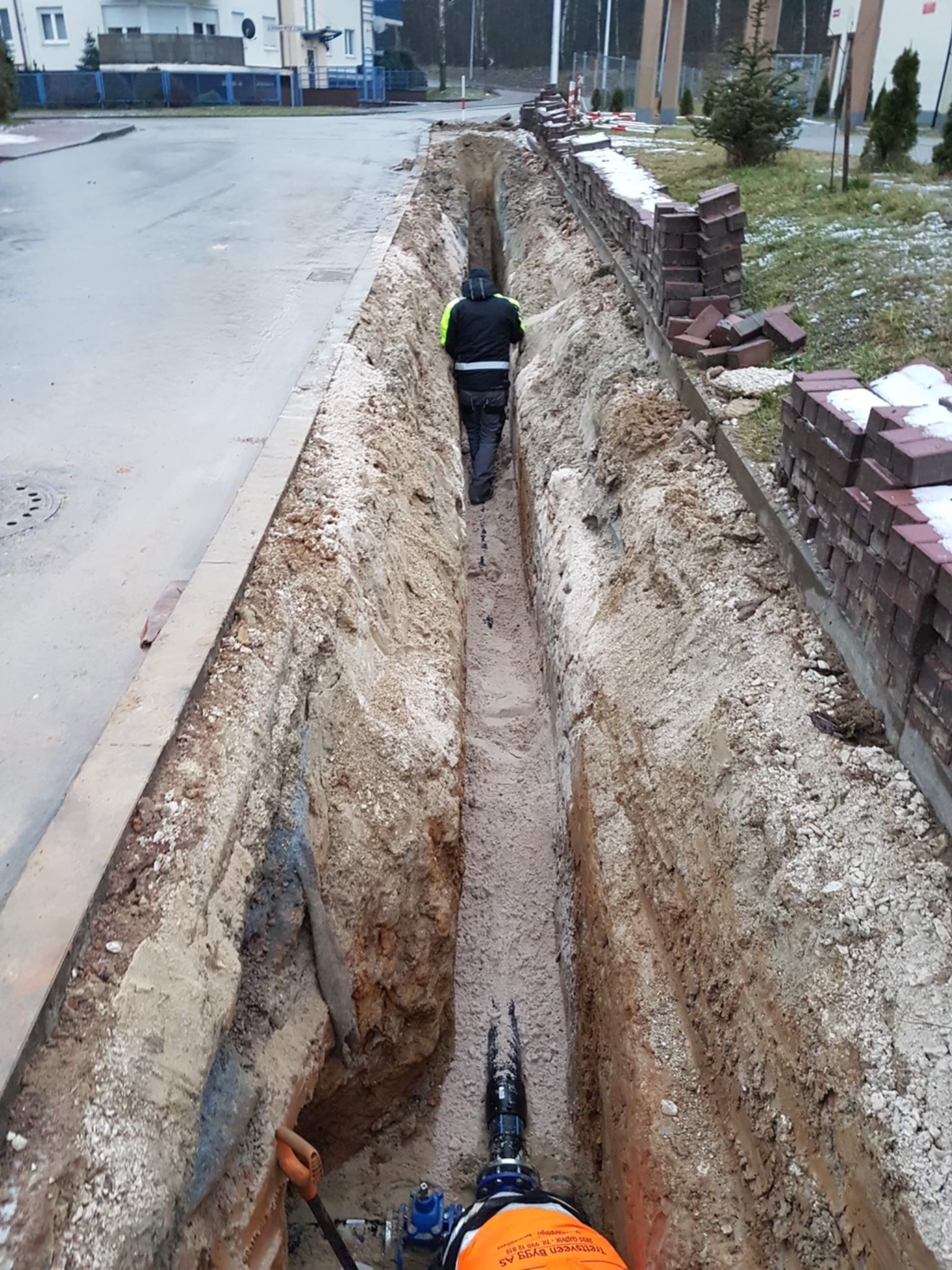 Wąski wykop w ziemi wzdłuż chodnika i muru z cegieł, pracownik w odblaskowej kamizelce wewnątrz wykopu, widoczne elementy instalacji wodociągowej i narzędzie w dole wykopu.