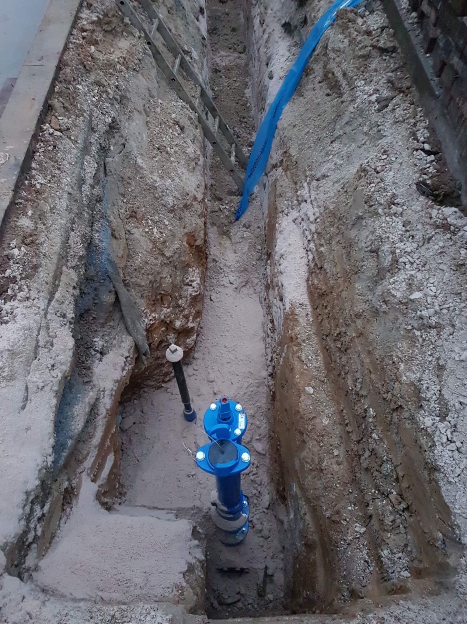 Wykop z zamontowanym niebieskim zaworem wodnym, widoczna drabina oparta o krawędź wykopu i fragment niebieskiej rury wodociągowej.