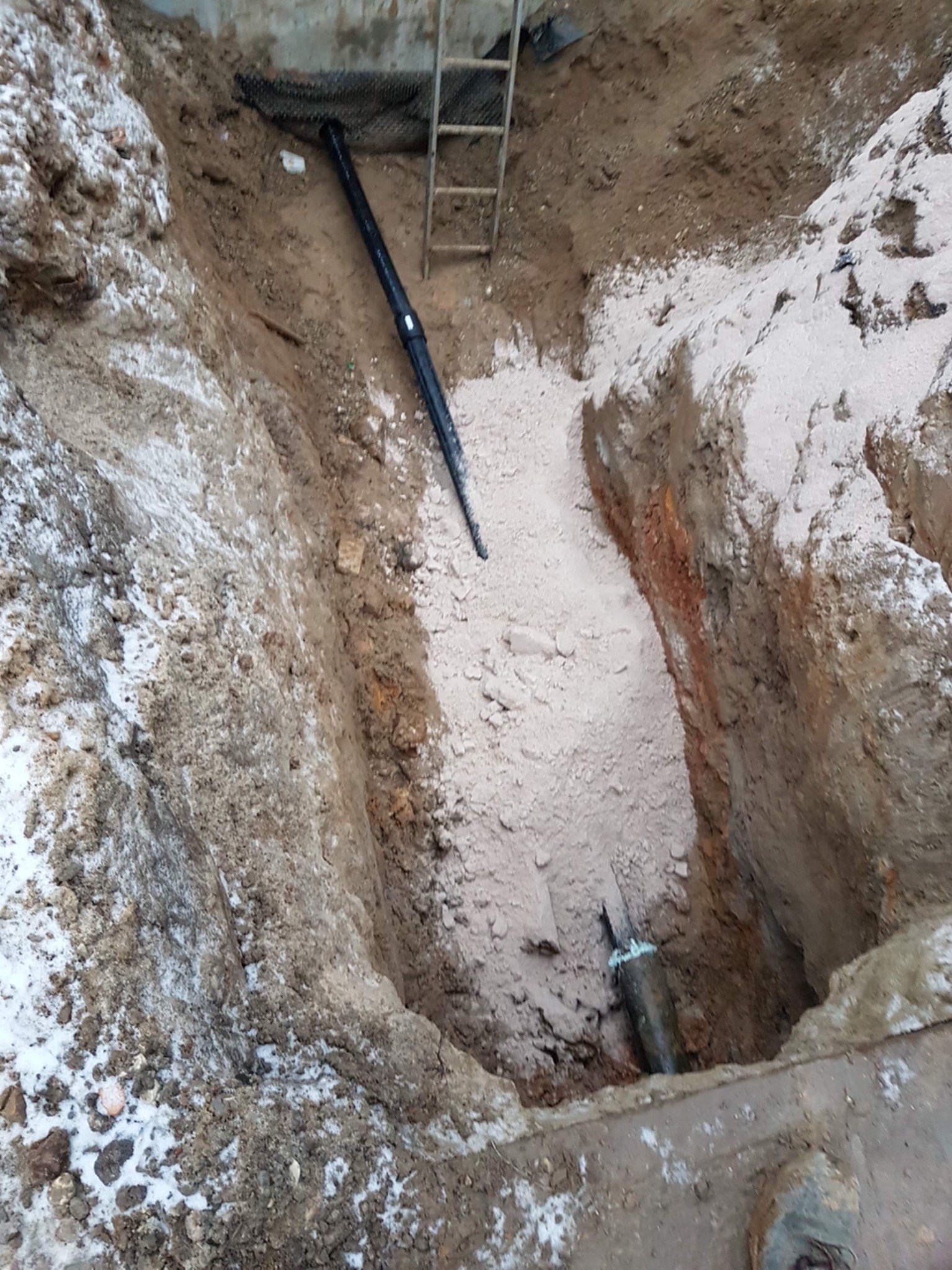 Wykop z widocznymi rurami kanalizacyjnymi, zasypany piaskiem, z fragmentami śniegu na krawędziach i drabiną w tle.