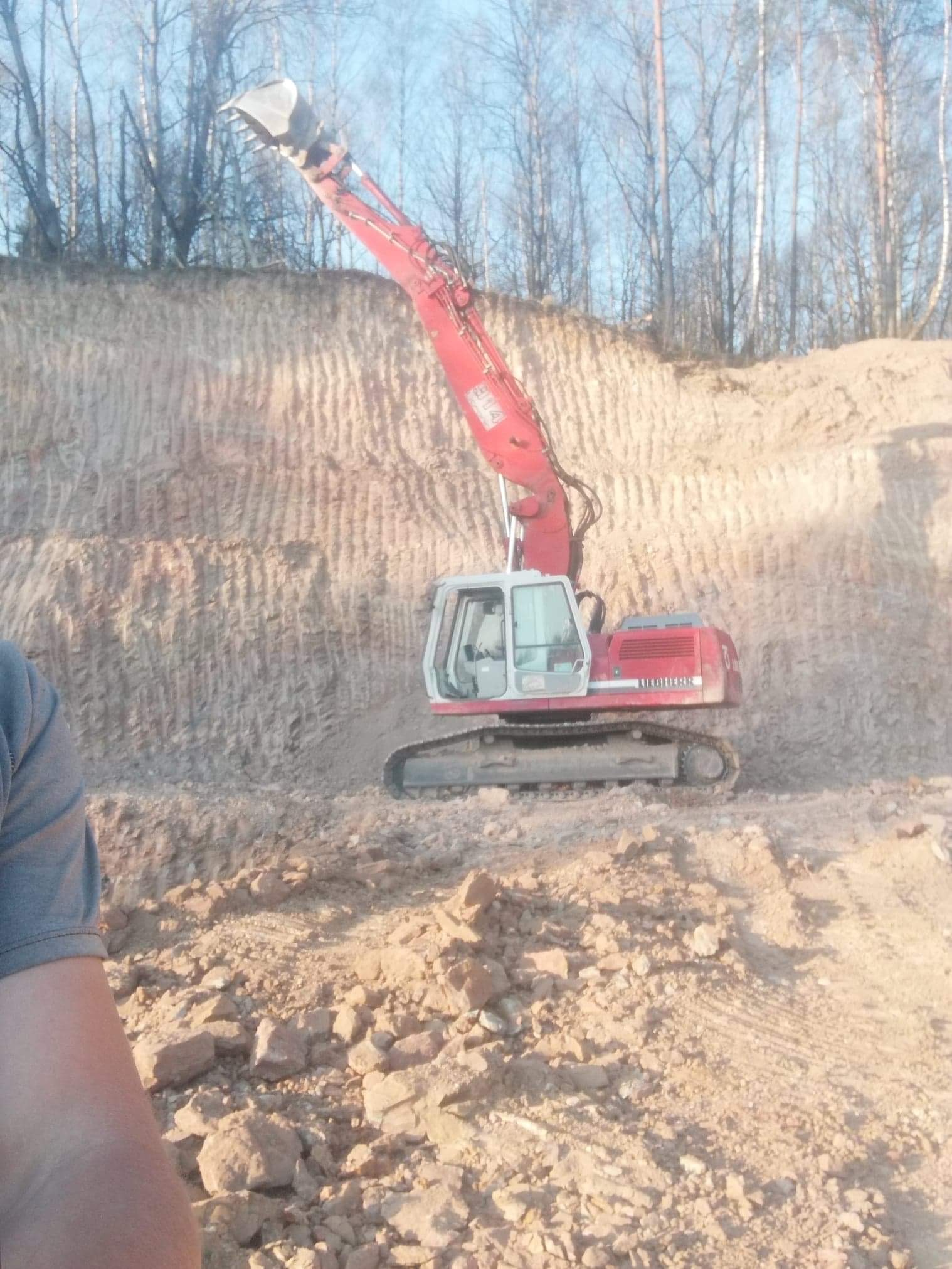 Czerwona koparka Liebherr na tle wykopu w ziemi, z widocznym fragmentem czyjegoś ramienia w lewym dolnym rogu kadru.