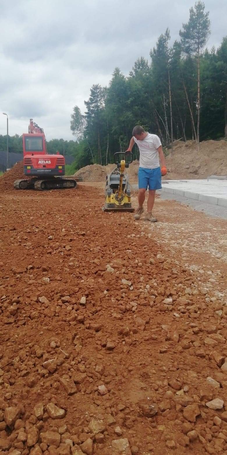 Pracownik w krótkich spodenkach obsługuje zagęszczarkę na placu budowy z czerwoną glebą, w tle koparka Atlas na usypanej ziemi i fragment betonowego fundamentu.