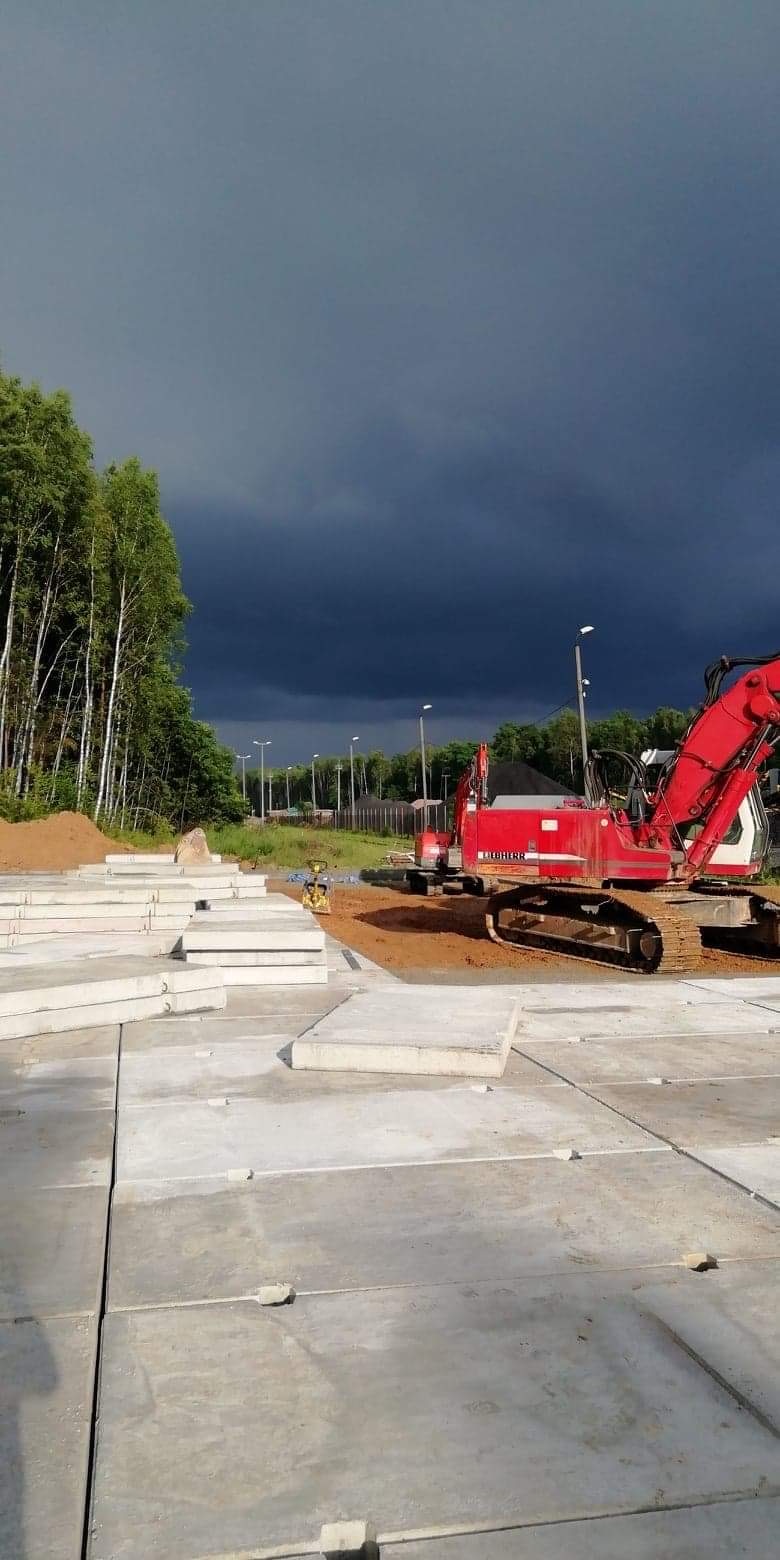 Czerwona koparka gąsienicowa na placu budowy z ułożonymi betonowymi płytami, na tle ciemnego, burzowego nieba i rzędu lamp ulicznych w oddali.