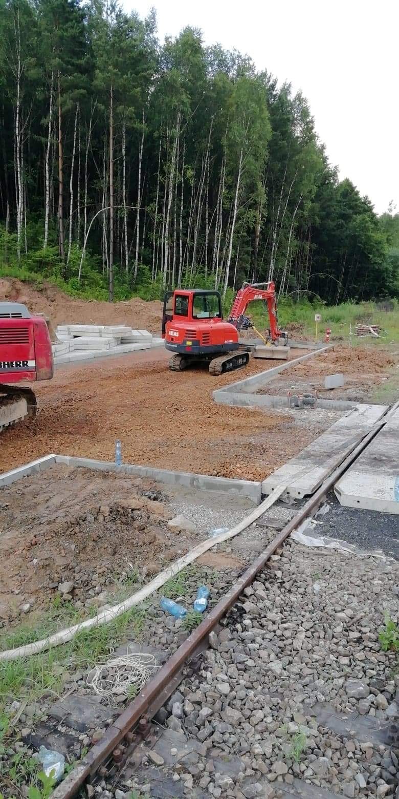 Czerwony mini-koparka marki Atlas na placu budowy z przygotowanym podłożem, betonowymi elementami i w tle las brzozowy.