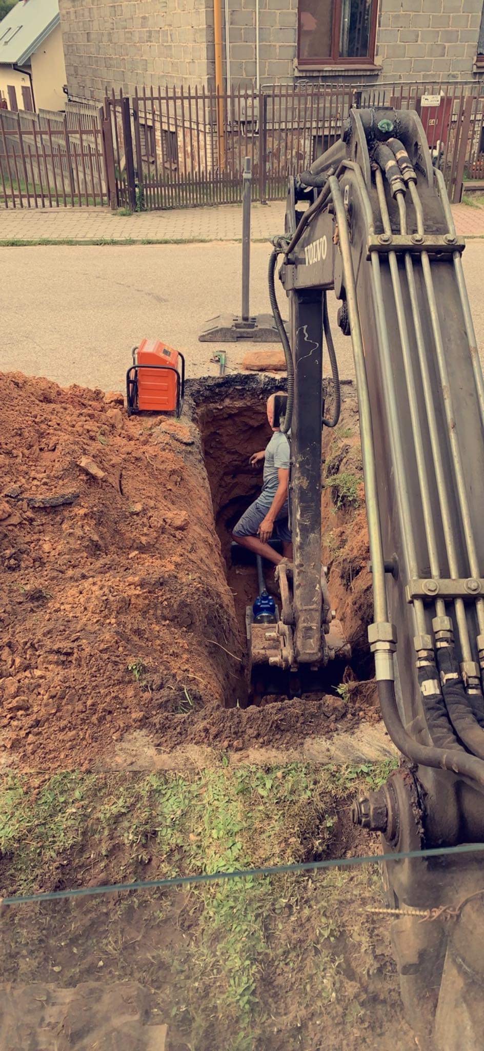 Operator koparki Volvo w wąskim wykopie ziemnym, obok stoi agregat prądotwórczy, widoczne instalacje wodne w dole wykopu, teren zabudowany w tle.