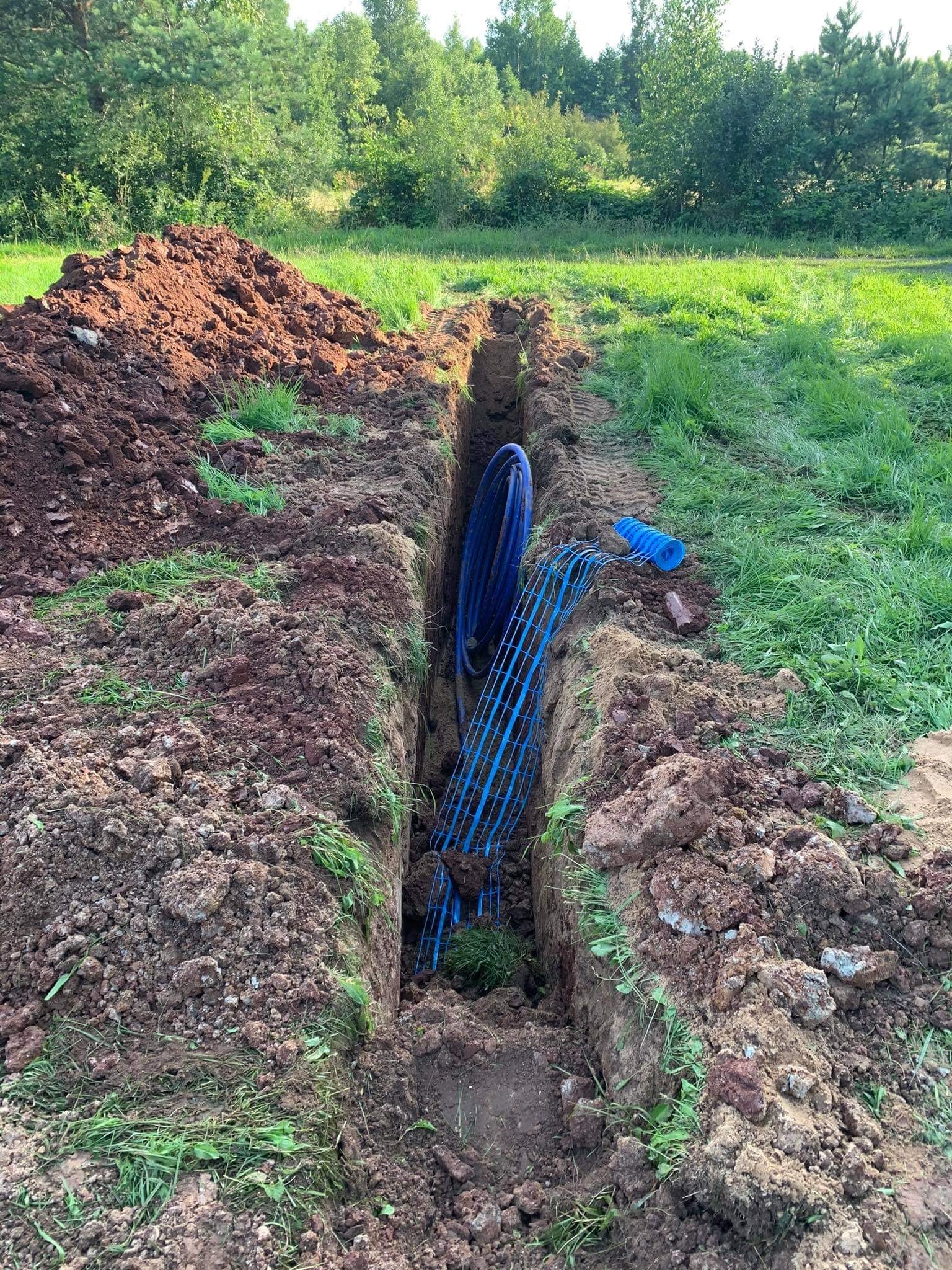 Wykop w ziemi na zewnątrz, z widocznymi niebieskimi rurami i siatką zabezpieczającą wewnątrz, obok wykopana ziemia i trawa.