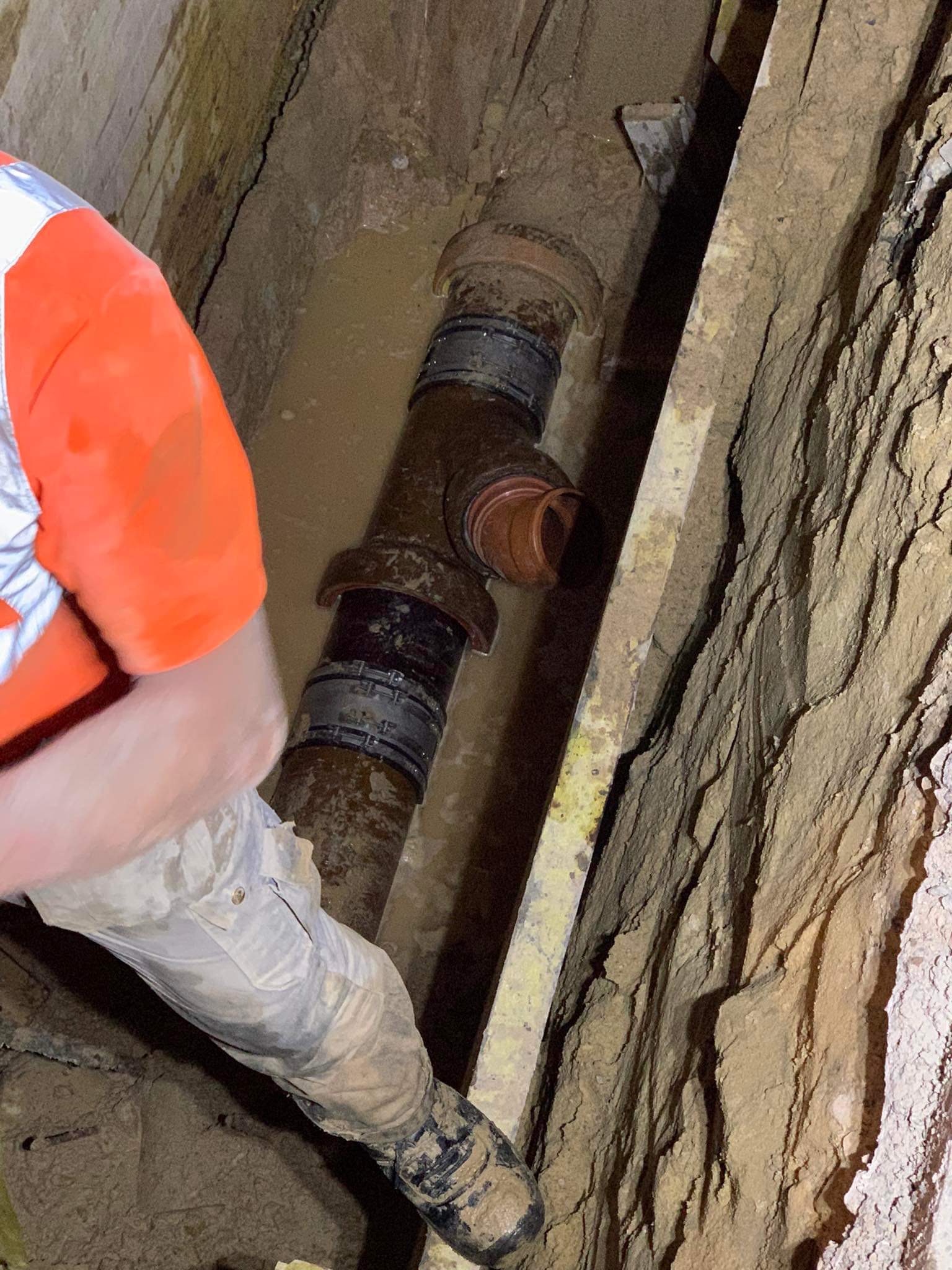 Pracownik w odblaskowej kamizelce i ubrudzonych spodniach w wykopie, obok rury kanalizacyjnej z łącznikami w błotnistym wykopie zabezpieczonym deskami.