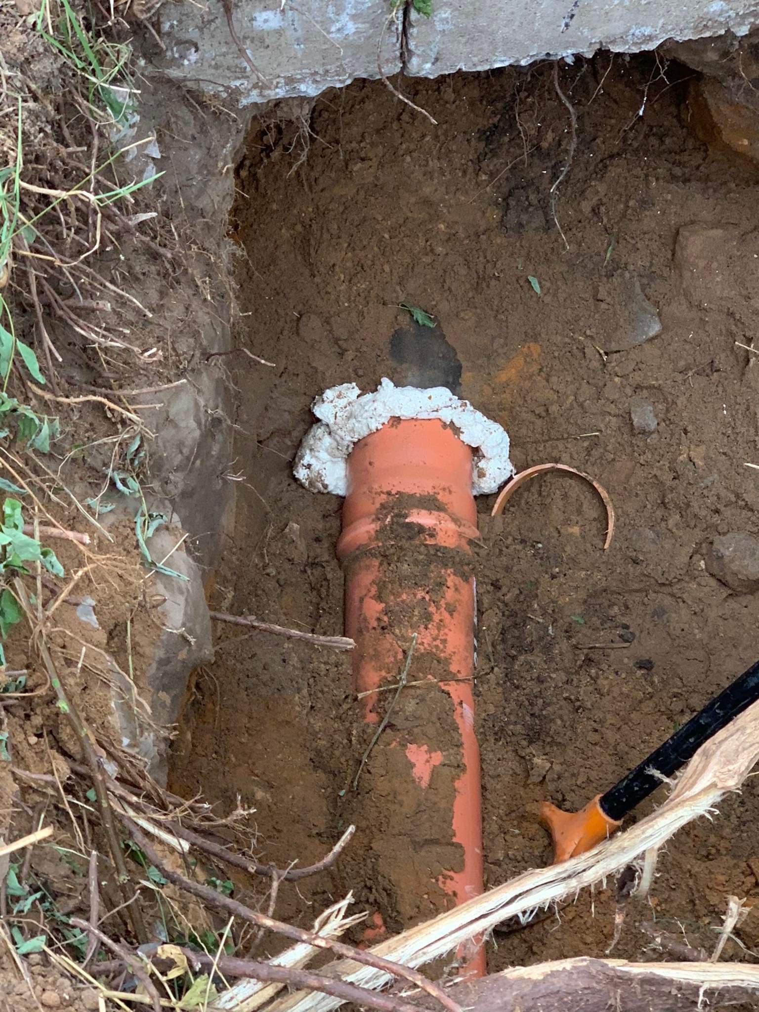 Pomarańczowa rura kanalizacyjna wystaje z wykopu w ziemi, uszczelniona pianką montażową przy betonowej krawędzi, widoczne narzędzie w prawym dolnym rogu.