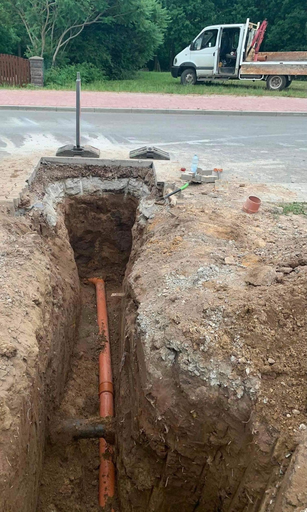 Wykop w ziemi z widocznymi pomarańczowymi rurami kanalizacyjnymi, na powierzchni ziemi leżą kostki brukowe i stoi biały samochód dostawczy