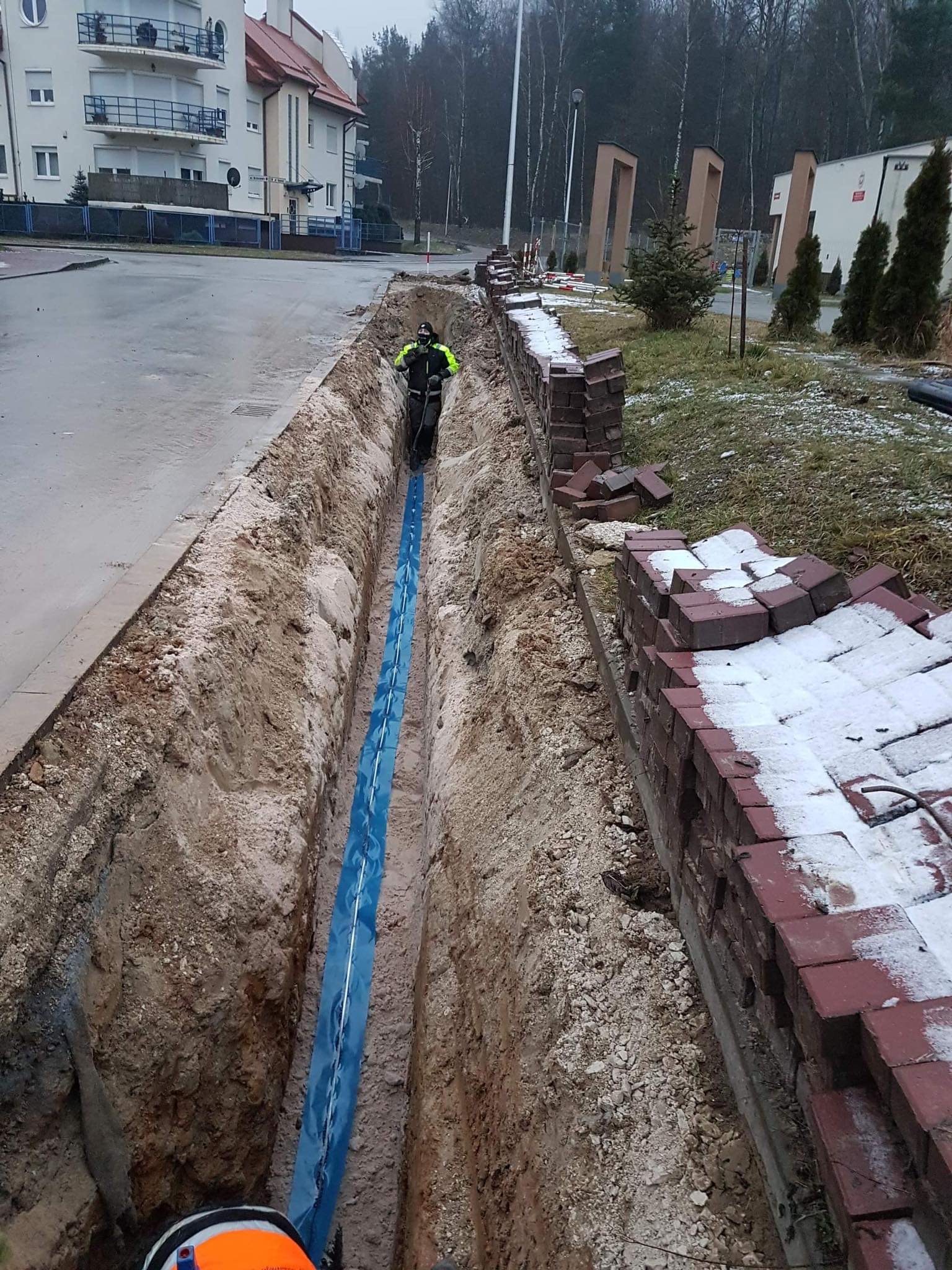 Wykop liniowy z ułożoną niebieską rurą w ochronnej folii, pracownik w odblaskowej kurtce w głębi wykopu, częściowo zdemontowany mur z cegły po prawej stronie, w tle budynki mieszkalne i drzewa.