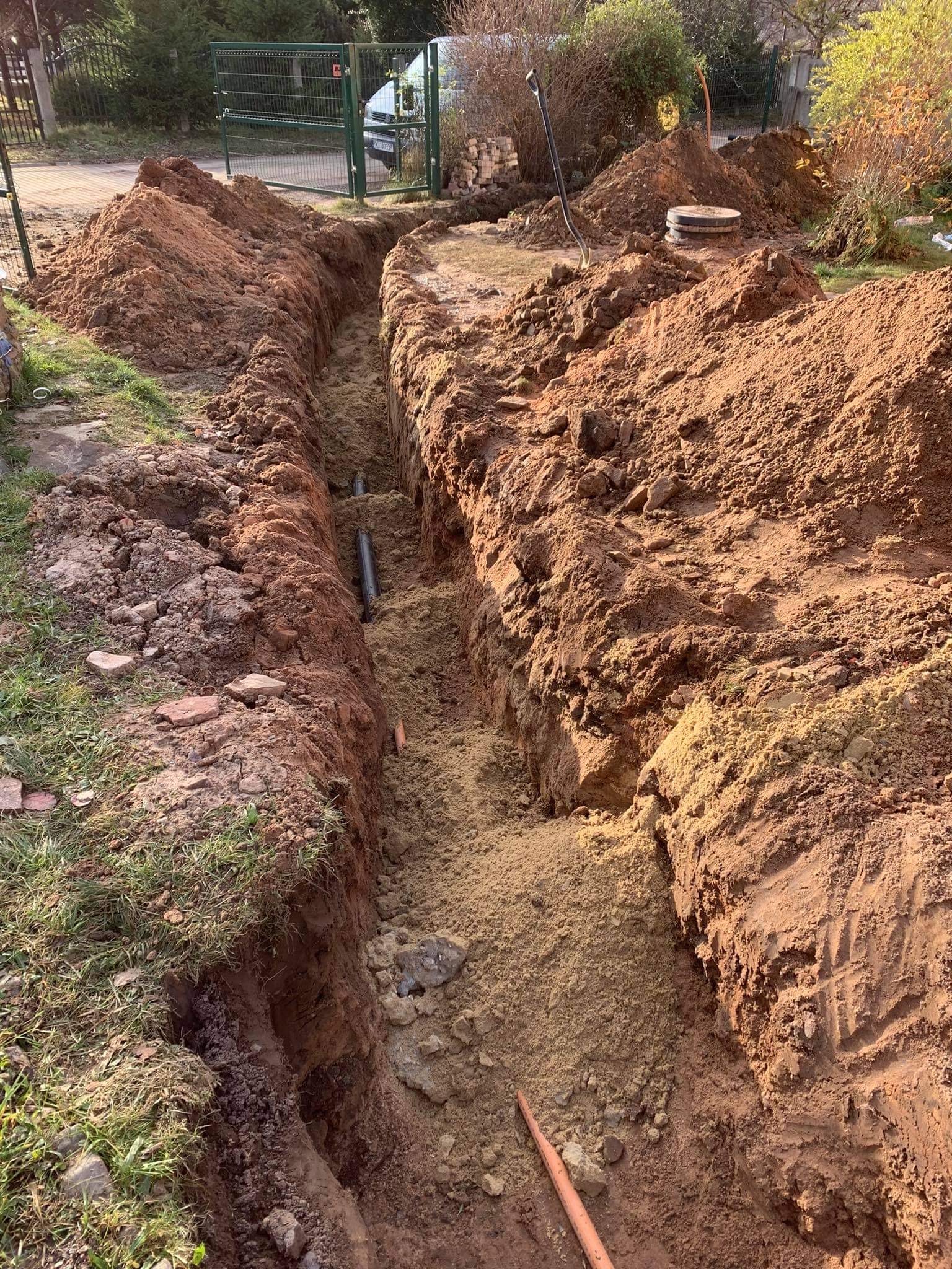 Wykopany rów w ziemi z widocznymi rurami kanalizacyjnymi, hałdy ziemi po obu stronach wykopu, teren ogrodzony siatką w tle.