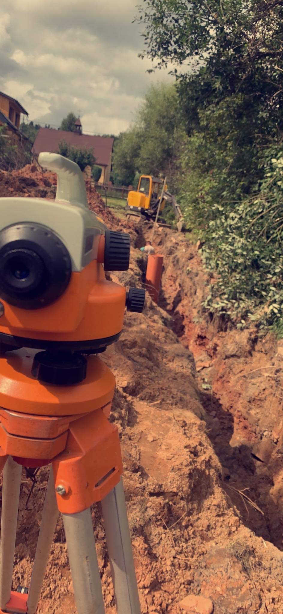 Pomarańczowy niwelator optyczny na trójnogu, w tle wykop w ziemi, rury kanalizacyjne i koparka, krajobraz wiejski.