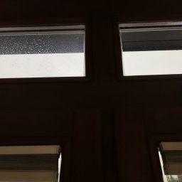 Czystomig - Okno z drewnianą ramą, górna lewa szyba pokryta kroplami wody, pozostałe czyste, dolne okna zasłonięte roletami.