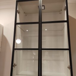 Czystomig - Pusta, czysta, biała szafka wisząca z przeszklonymi drzwiami w czarnej ramie, oświetlona od wewnątrz, zamontowana na ścianie.