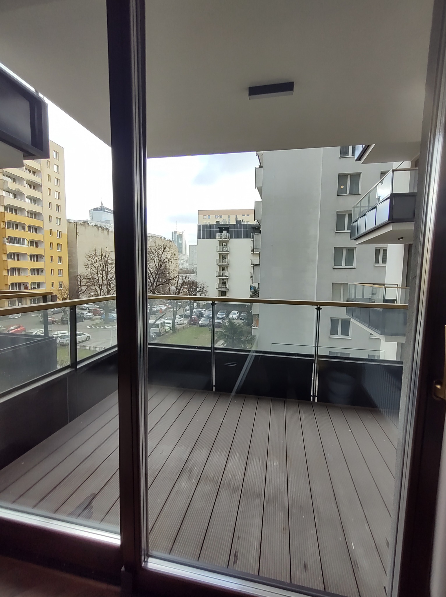 Balkon z drewnianą podłogą i szklaną balustradą, widok na parking i budynki mieszkalne w tle.