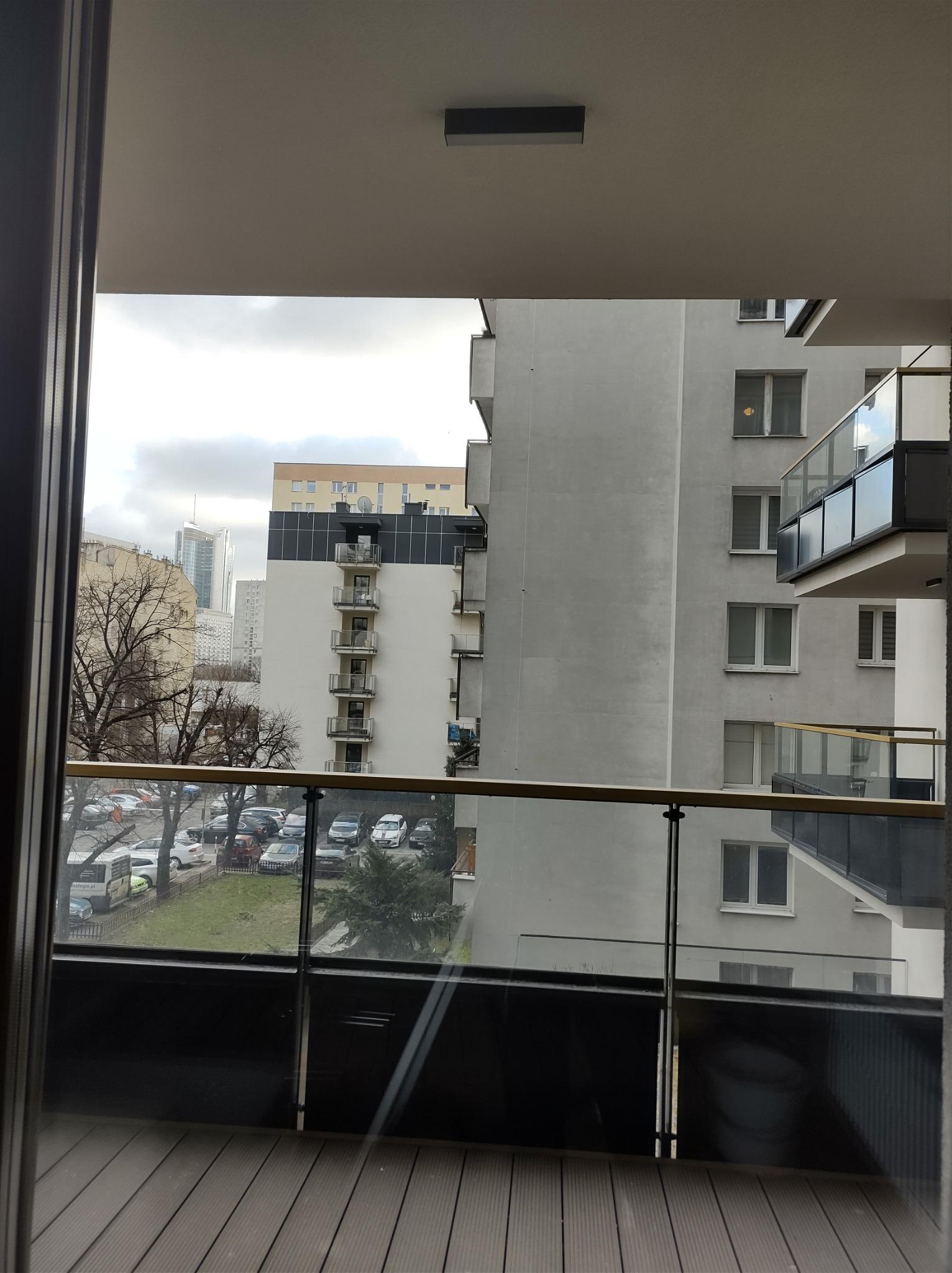 Widok z balkonu na osiedle mieszkaniowe w miejskiej scenerii, z widocznymi drzewami bez liści, zaparkowanymi samochodami i budynkami o różnej wysokości.
