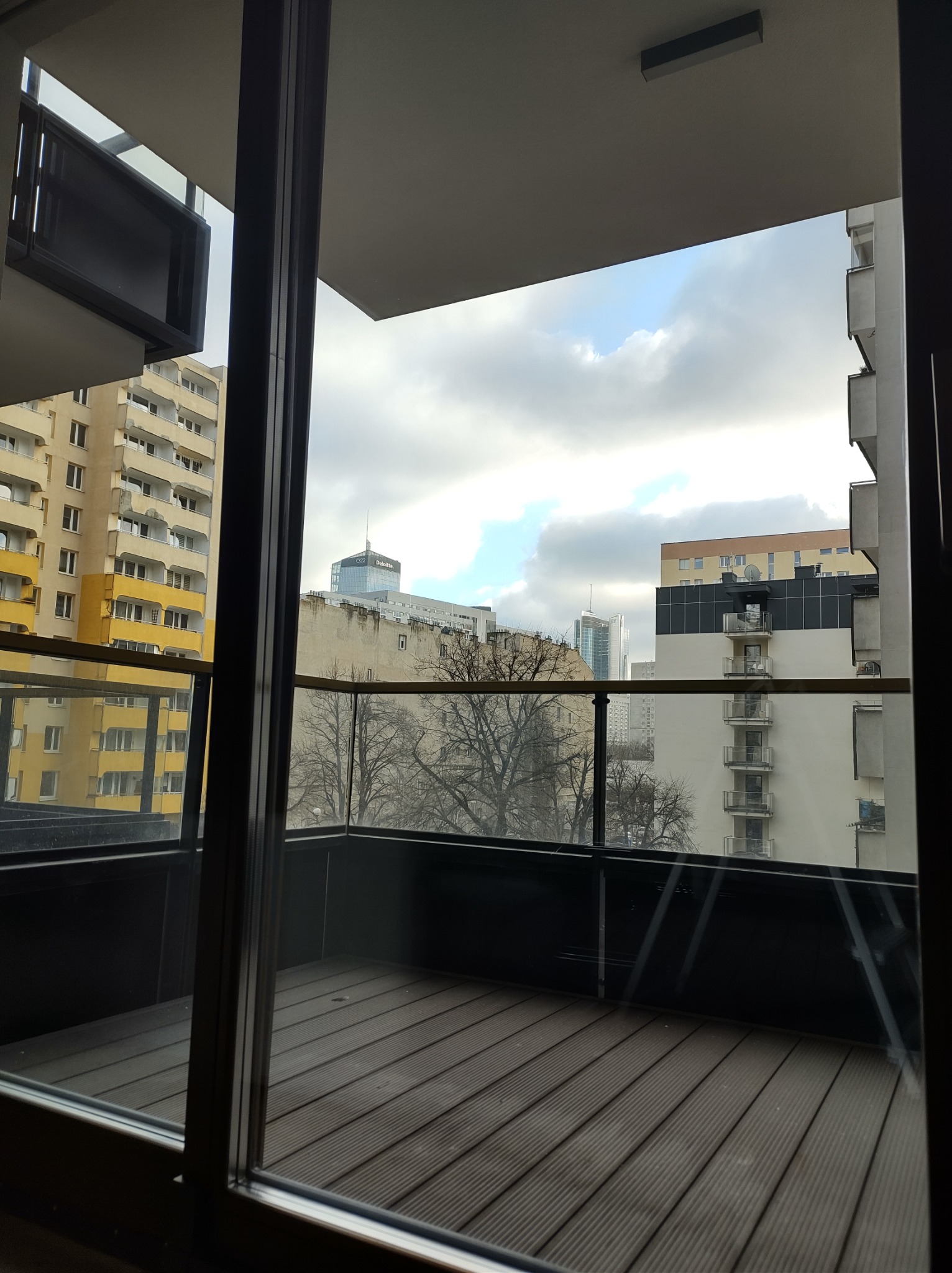 Widok z balkonu w Warszawie na budynki mieszkalne i biurowce w pochmurny dzień. Widoczne drewniane deski podłogowe na balkonie oraz szklana balustrada.