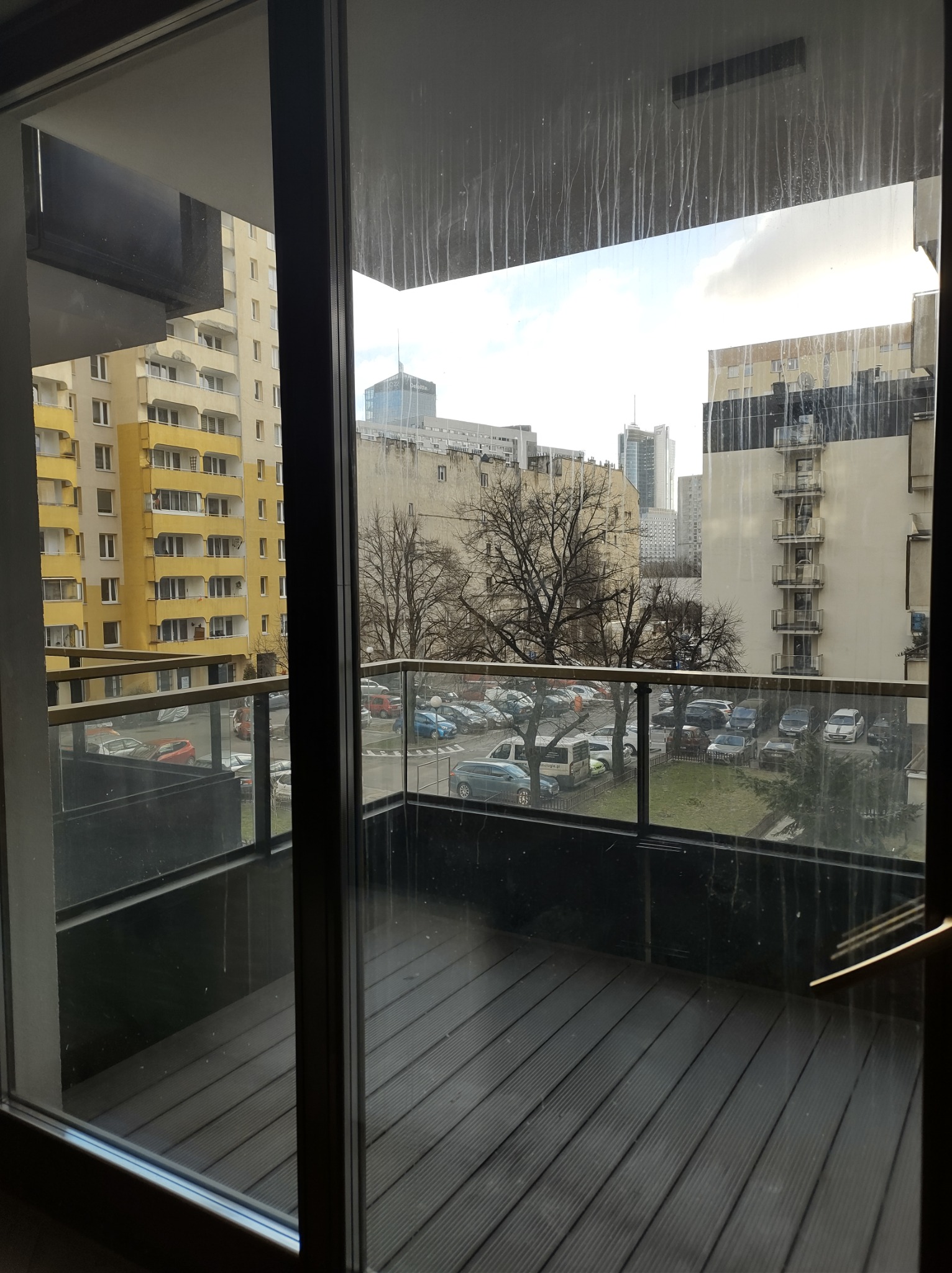 Widok przez brudne okno balkonowe na panoramę miasta z parkingiem i budynkami w tle. Na szybie widoczne zacieki.