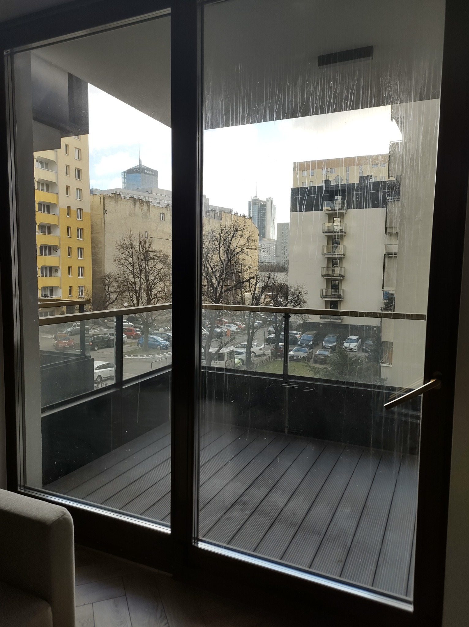 Widok z balkonu przez brudne, przesuwne okno balkonowe na panoramę miasta z budynkami mieszkalnymi i biurowcami w tle.